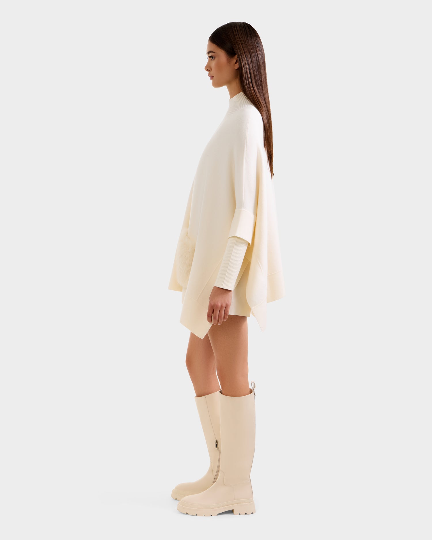 A woman in profile wears the Rudsak SWOON - 8525672 Vanilla sweater poncho with faux fur pockets, a matching long-sleeve top, and knee-high cream boots. Her straight brown hair is down against a plain white background.