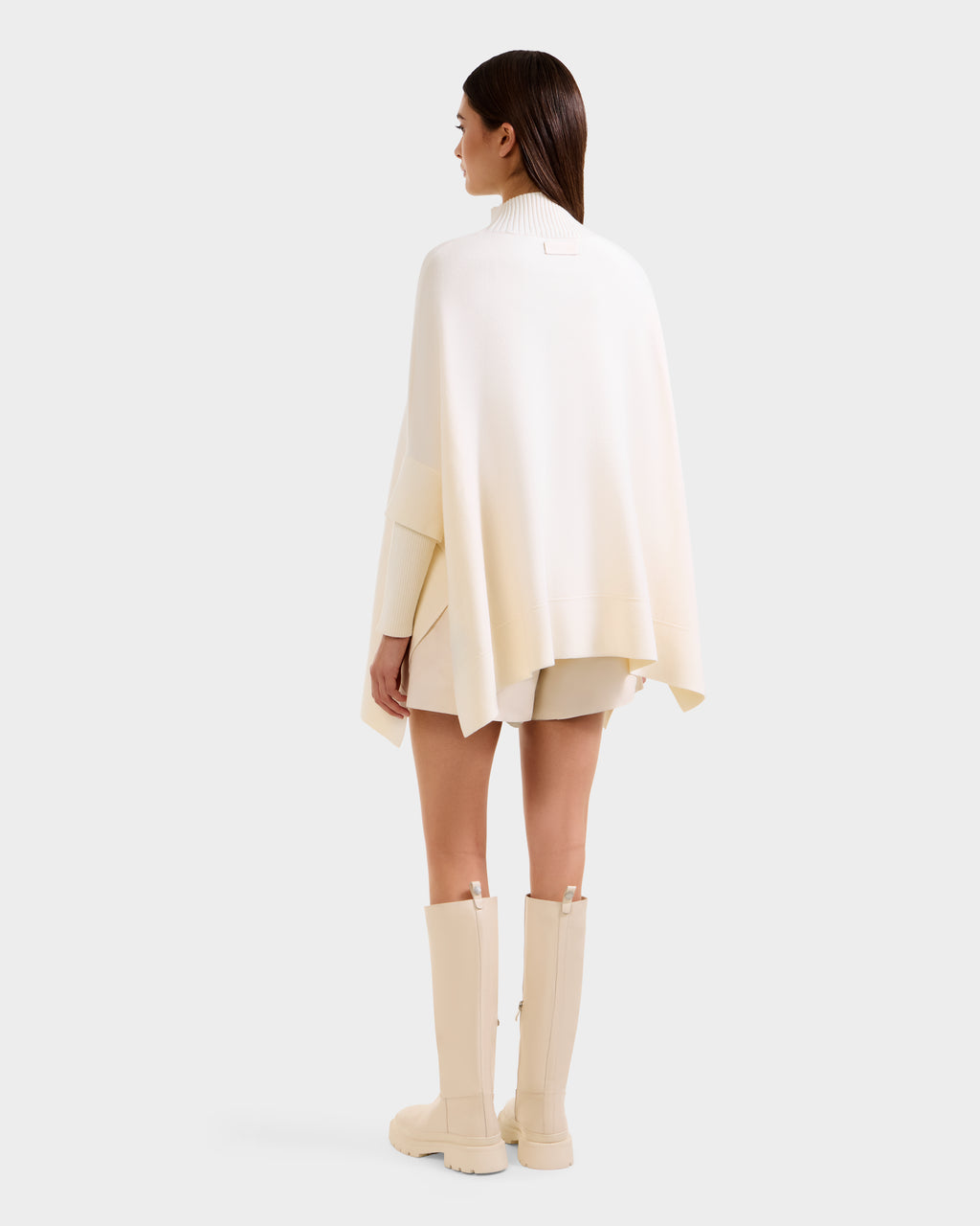 A woman faces away, modeling the Rudsak SWOON - 8525672 Vanilla sweater poncho with matching shorts and beige knee-high boots. Her straight brown hair is worn down, set against a plain white background.