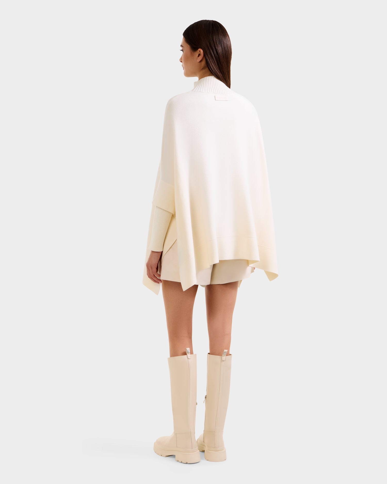 A woman faces away, modeling the Rudsak SWOON - 8525672 Vanilla sweater poncho with matching shorts and beige knee-high boots. Her straight brown hair is worn down, set against a plain white background.