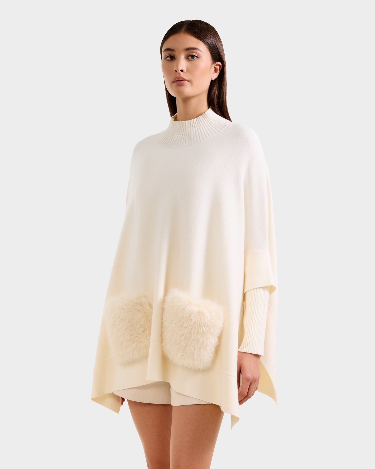 Wearing the SWOON - 8525672 Vanilla sweater poncho by Rudsak, featuring a ribbed high neckline and faux fur pockets, paired with matching shorts, a woman stands against a plain background—showcasing chic modern layering.