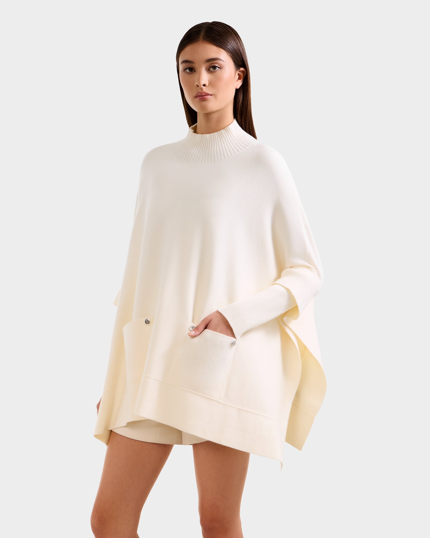 A woman models the Rudsak SWOON - 8525672 Vanilla, a loose ivory sweater poncho with a high ribbed collar and large front pockets, paired with matching shorts, standing against a light gray background.
