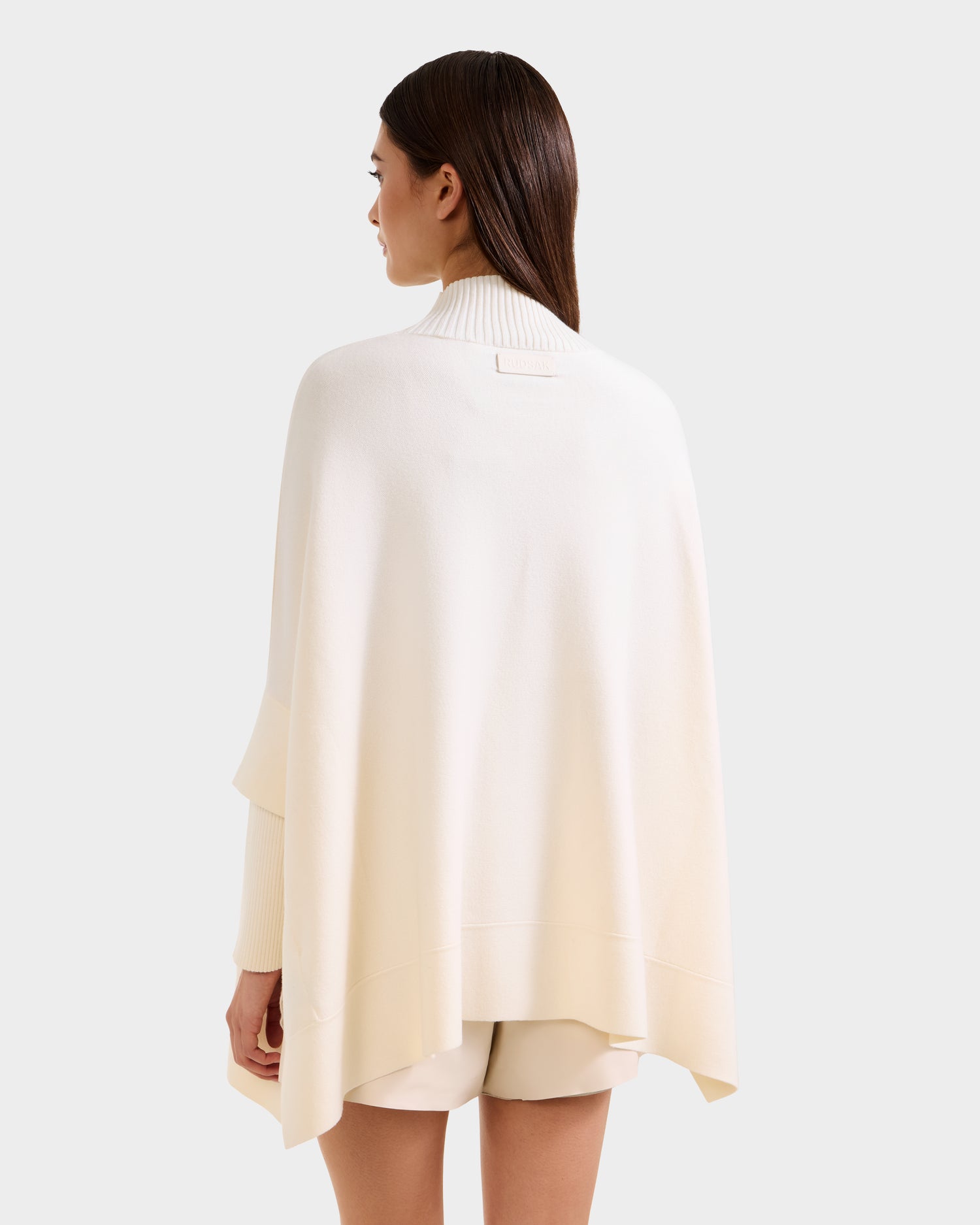 A woman with straight brown hair, seen from behind, wears Rudsak’s SWOON - 8525672 Vanilla: a modern cream sweater poncho with ribbed turtleneck and matching cream shorts, set against a plain light background.