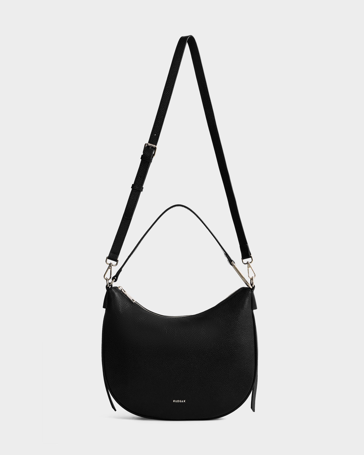 The Rudsak SYLVIA - L325515 Black is a women's leather crescent-shaped shoulder bag with an adjustable long strap, short handle, gold hardware, and a subtle embossed logo on the front, shown against a white background.