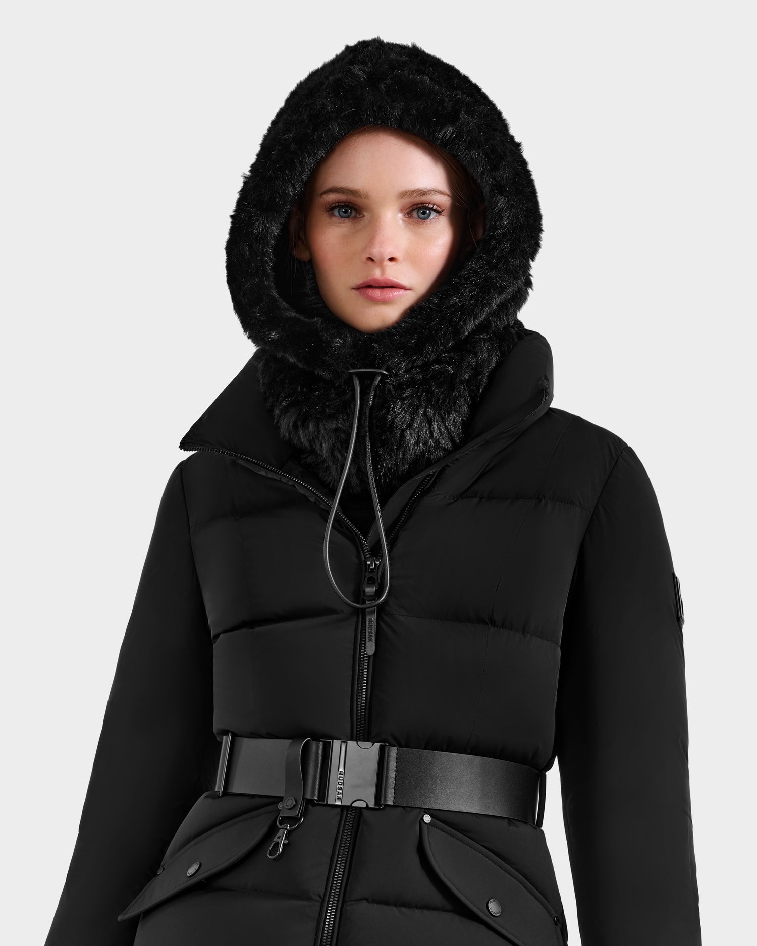 A woman models the Rudsak TARA - 8425025 Black puffer coat with a fur-lined hood and wide belt, standing against a plain light background and exuding chic winter style as she looks directly at the camera.