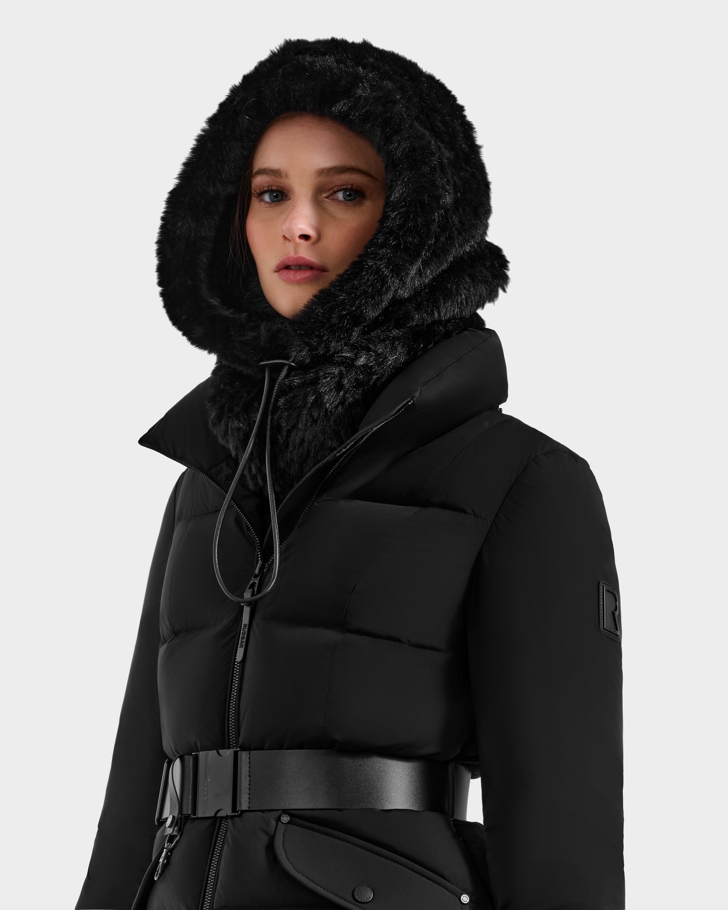 A woman models the Rudsak TARA - 8425025 Black puffer coat with a faux fur hood and wide belt, styled with a women’s knitted faux fur balaclava, elevating her winter look against a plain light background.