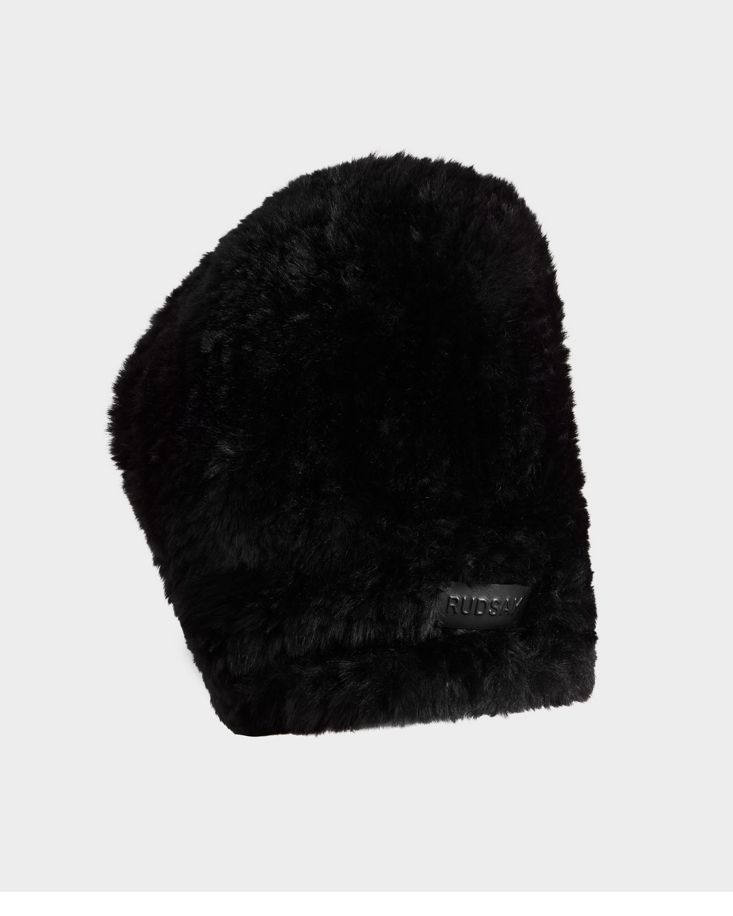 The TARA - 8425025 Black by Rudsak is a black faux fur winter hat with a slightly curved design and a small rectangular RUSSIA tag. This cozy accessory adds winter style, shown here against a plain white background.