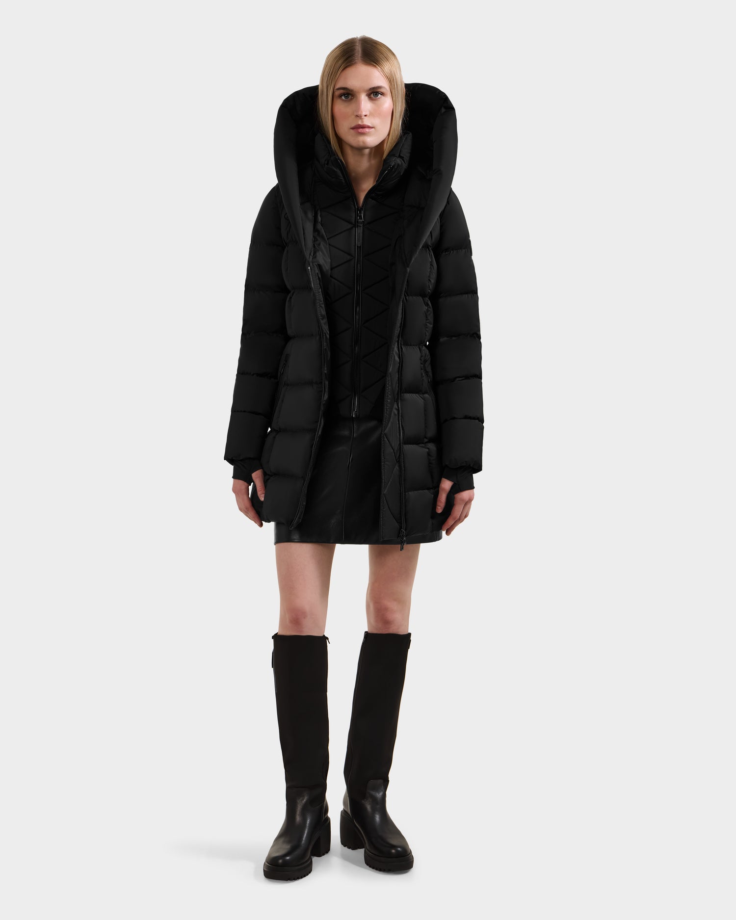 A woman faces forward in a Rudsak TAYLOR M - 8124545 Black water-repellent down puffer jacket with a large hood, styled over a black dress and knee-high boots. She stands against a plain white background.