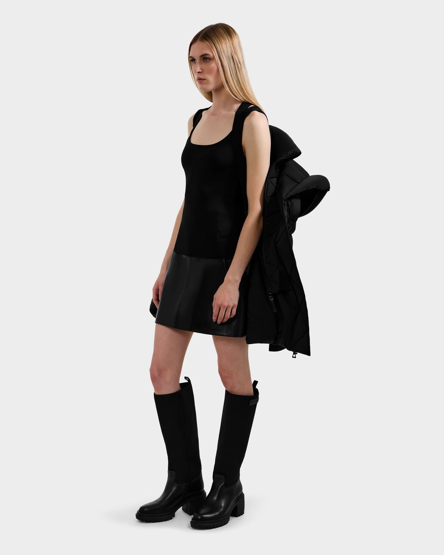 A woman with straight blonde hair poses against a light gray background, wearing the Rudsak TAYLOR M - 8124545 Black water-repellent jacket draped over her shoulder, paired with a black sleeveless top, short skirt, and knee-high boots.