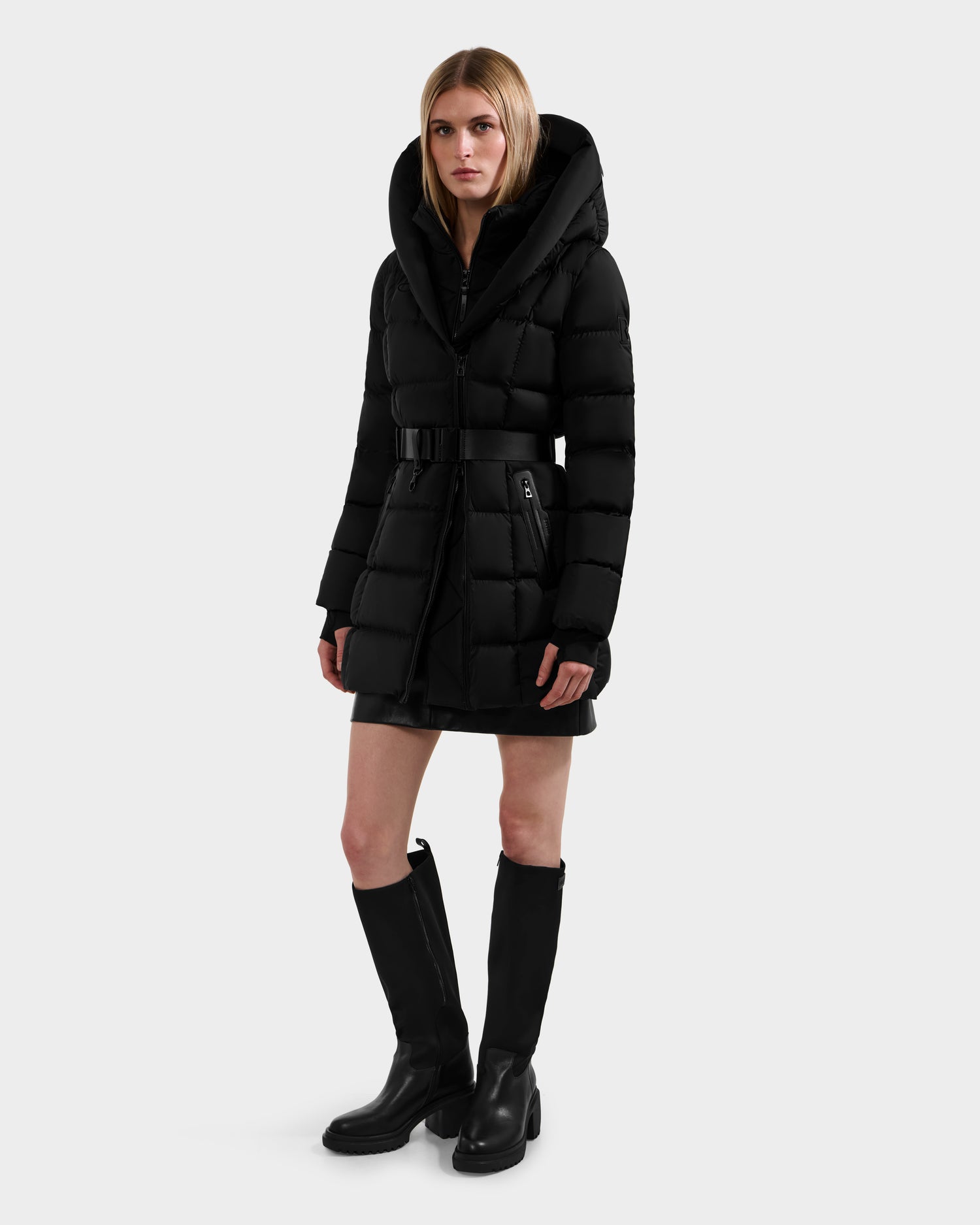 A woman with straight blonde hair and a neutral expression wears the Rudsak TAYLOR M - 8124545 Black water-repellent women's down puffer jacket with hood and waist belt, styled with black knee-high boots against a plain light gray background.