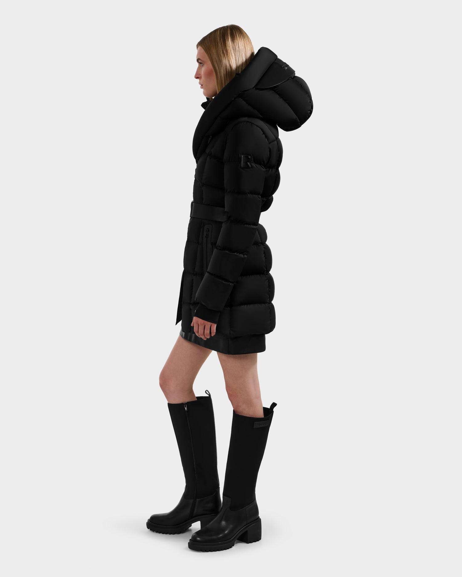 A woman in the Rudsak TAYLOR M - 8124545 black water-repellent winter jacket with a large hood and knee-high black boots stands in profile against a plain light gray background.