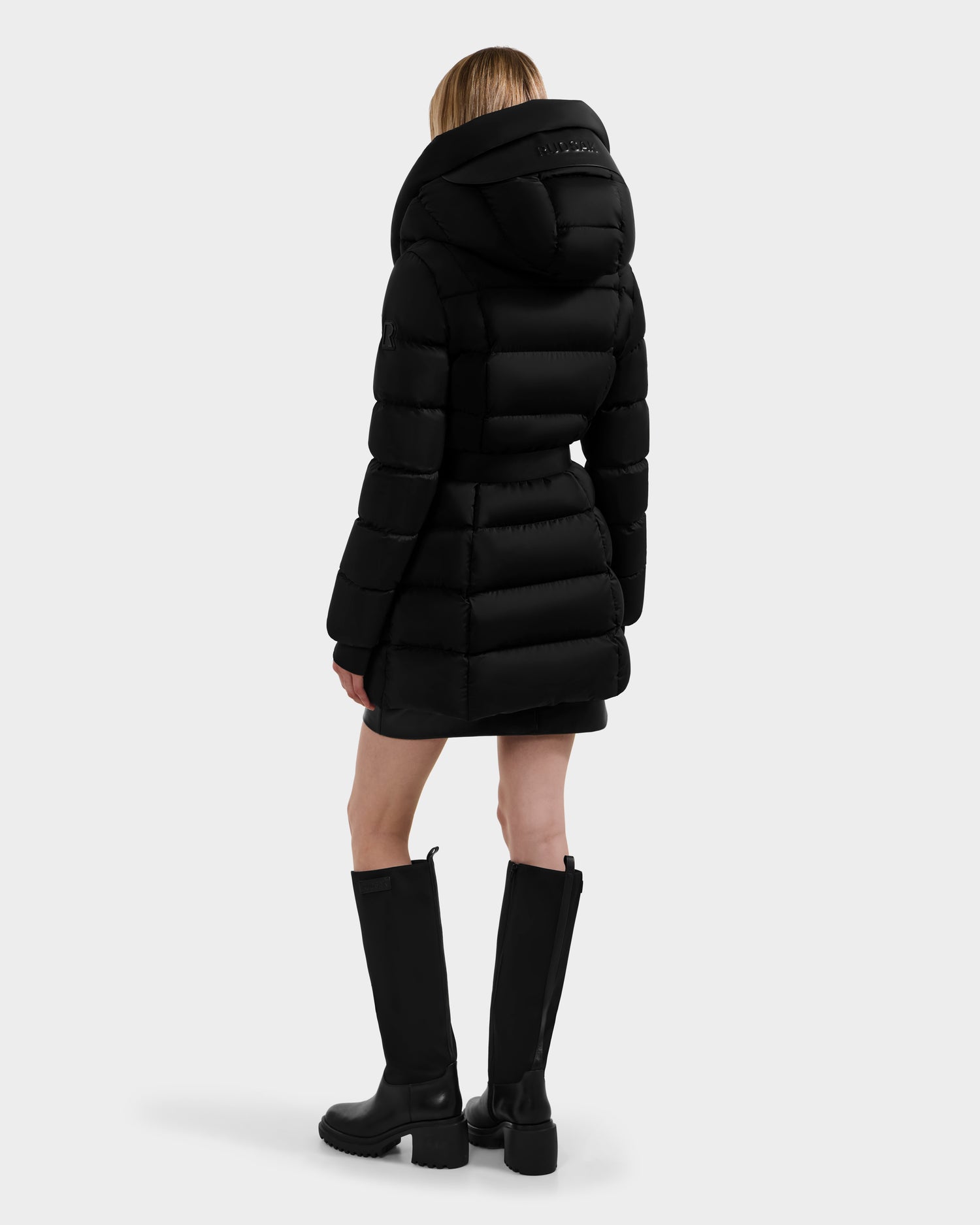 A person with light hair wears the Rudsak TAYLOR M - 8124545 Black, a long women’s down puffer jacket with a large hood and water-repellent finish, paired with knee-high black boots against a plain, light background.