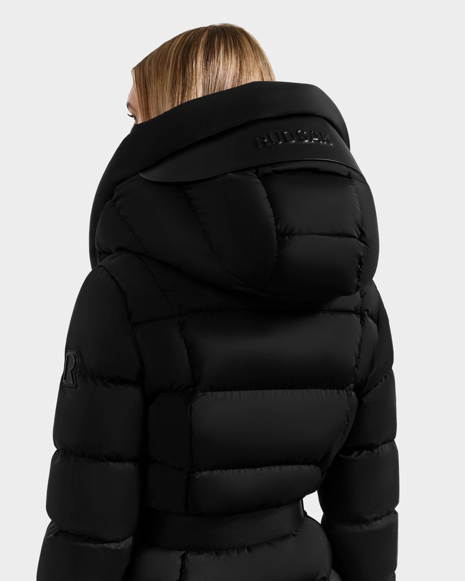 A blonde woman is shown from the back wearing the Rudsak TAYLOR M - 8124545 Black down puffer jacket, featuring a large high collar, hood, visible quilting, and logo detailing on the left sleeve and back of the hood.