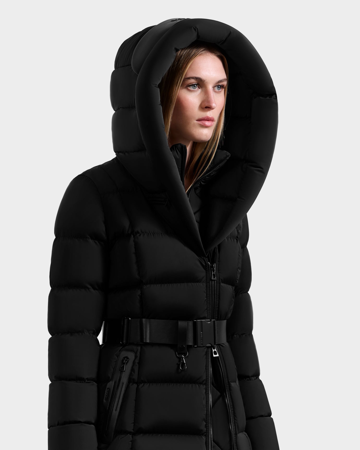 A woman poses against a plain light background wearing the Rudsak TAYLOR M - 8124545, a black belted quilted winter jacket with a large hood.