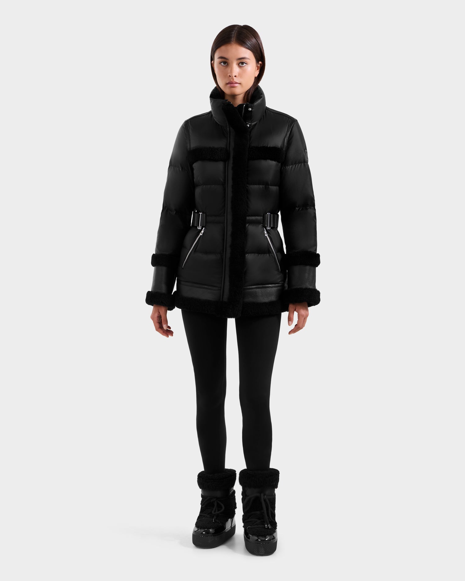 A woman stands facing forward in the Rudsak TESSA - 8125615 Black luxury winter coat, a belted black puffer jacket, paired with black leggings and winter boots against a plain light background.