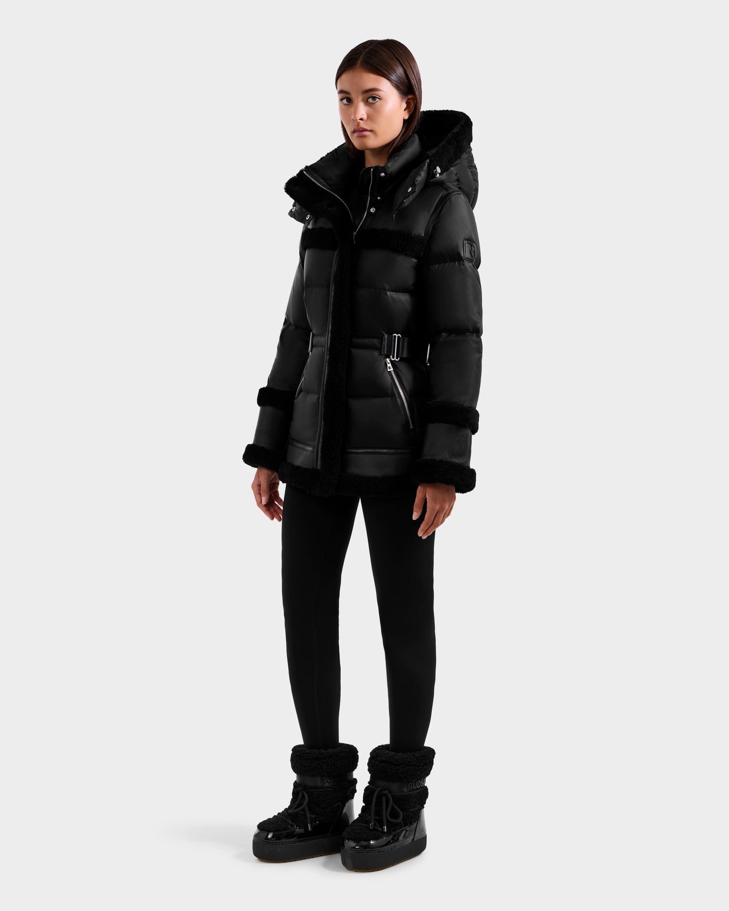 A person stands facing forward in the Rudsak TESSA - 8125615 Black women’s down puffer jacket with a belt, paired with black pants and shearling-trimmed black boots against a plain white background.