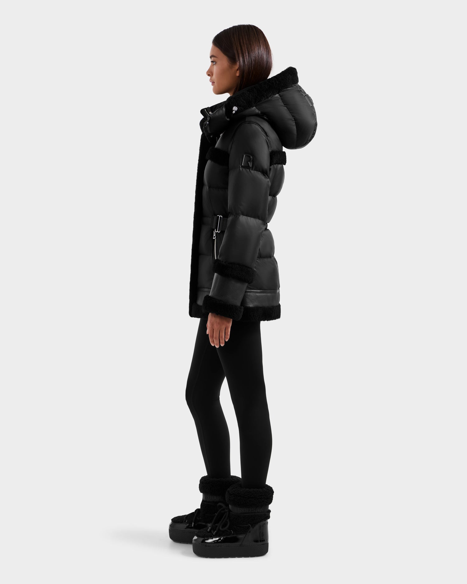 A woman in profile wears the Rudsak TESSA - 8125615 Black women's down puffer with hood, paired with black leggings and black winter boots, set against a plain light background.