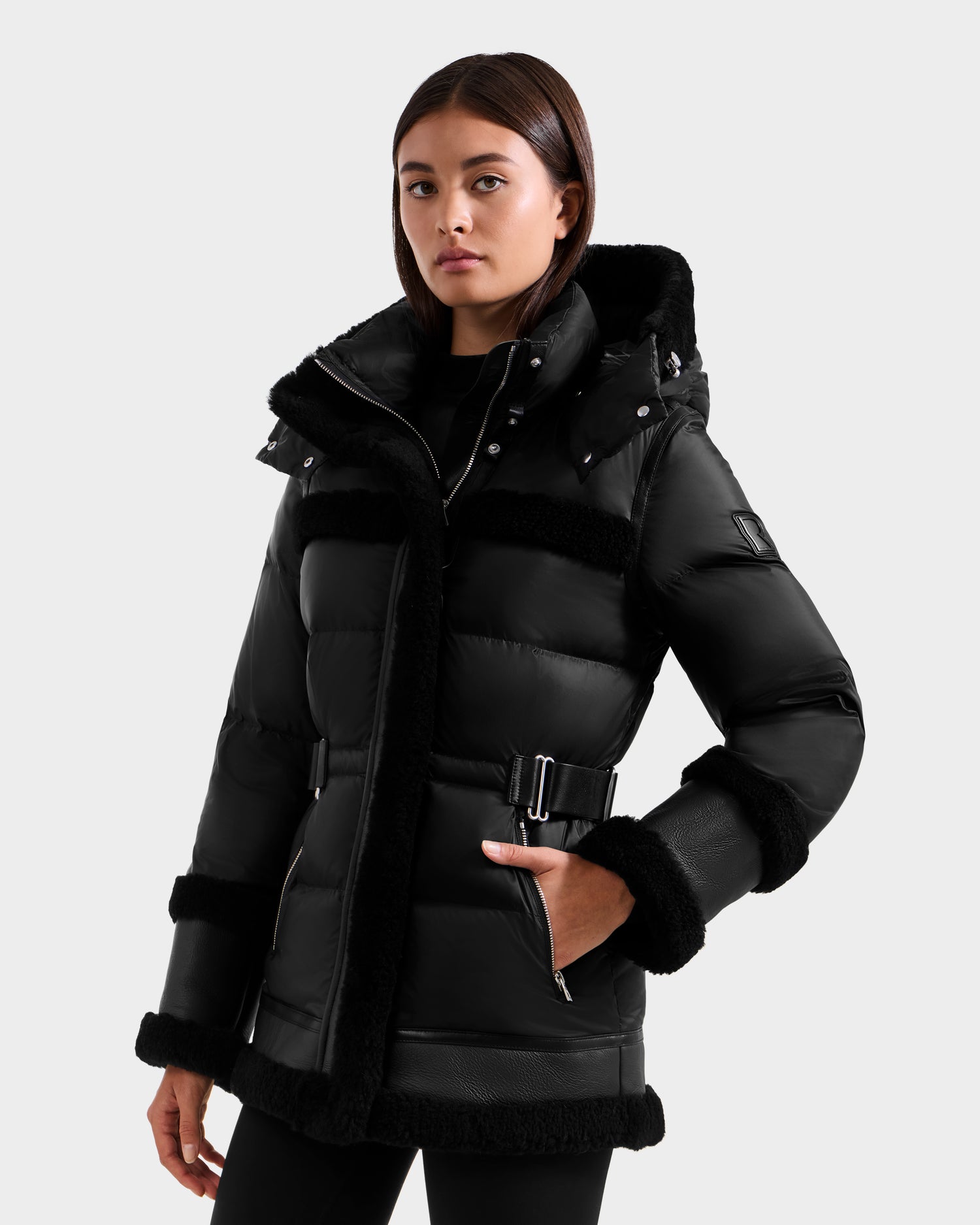A woman in the Rudsak TESSA - 8125615 Black down puffer jacket with shearling trim and belt stands against a plain light background, facing slightly to the side with a neutral expression.