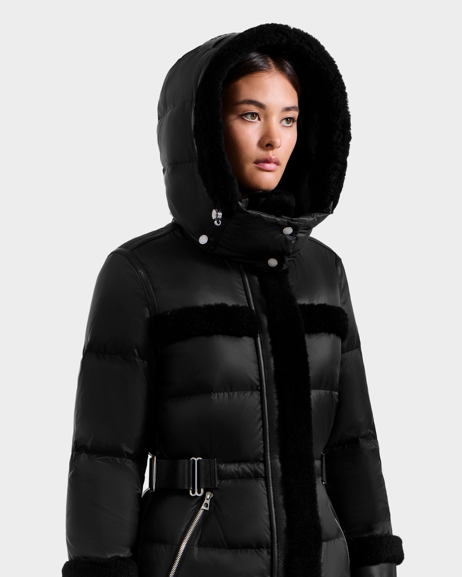A woman models the Rudsak TESSA - 8125615 Black, a luxury women's down puffer coat with a large hood, shearling trim, and zippered pockets, against a plain white background while looking slightly to the side.
