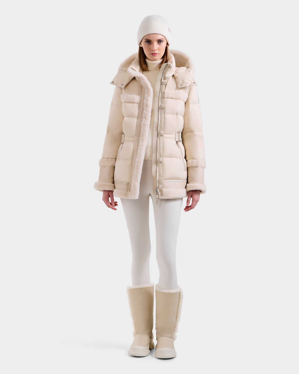 A woman models the Rudsak TESSA - 8125615 Vanilla, an off-white women's down puffer jacket with shearling trim, styled with white leggings, fur-lined boots, a white turtleneck, and a white knit beanie against a plain backdrop.