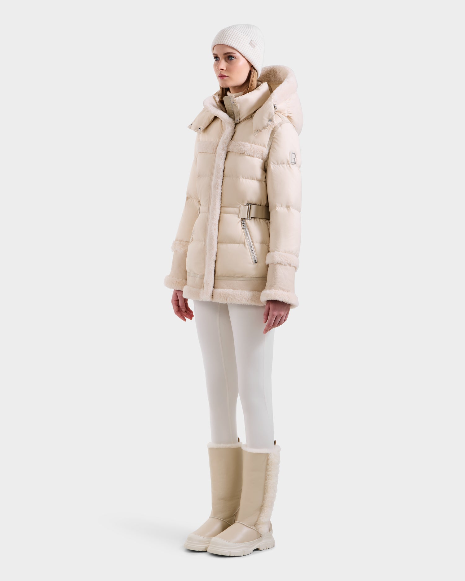 A woman wears the Rudsak TESSA - 8125615 Vanilla down puffer jacket with shearling trim, paired with white leggings, matching boots, and a white knit beanie, standing against a plain white background.