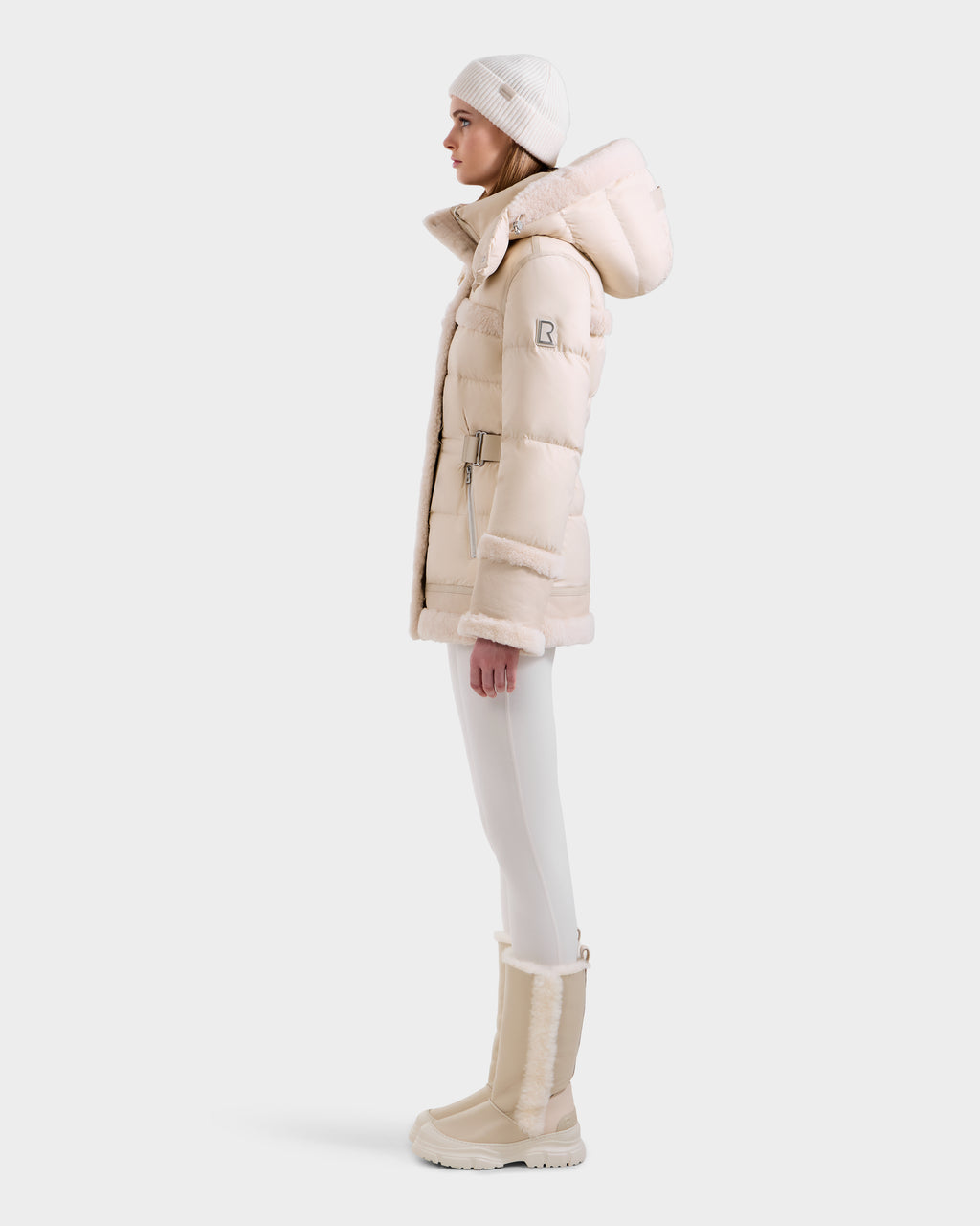 A woman in profile wears the Rudsak TESSA - 8125615 Vanilla winter jacket with shearling trim, styled with white pants, fur-trimmed boots, and a knit hat against a light background—a true winter essential.