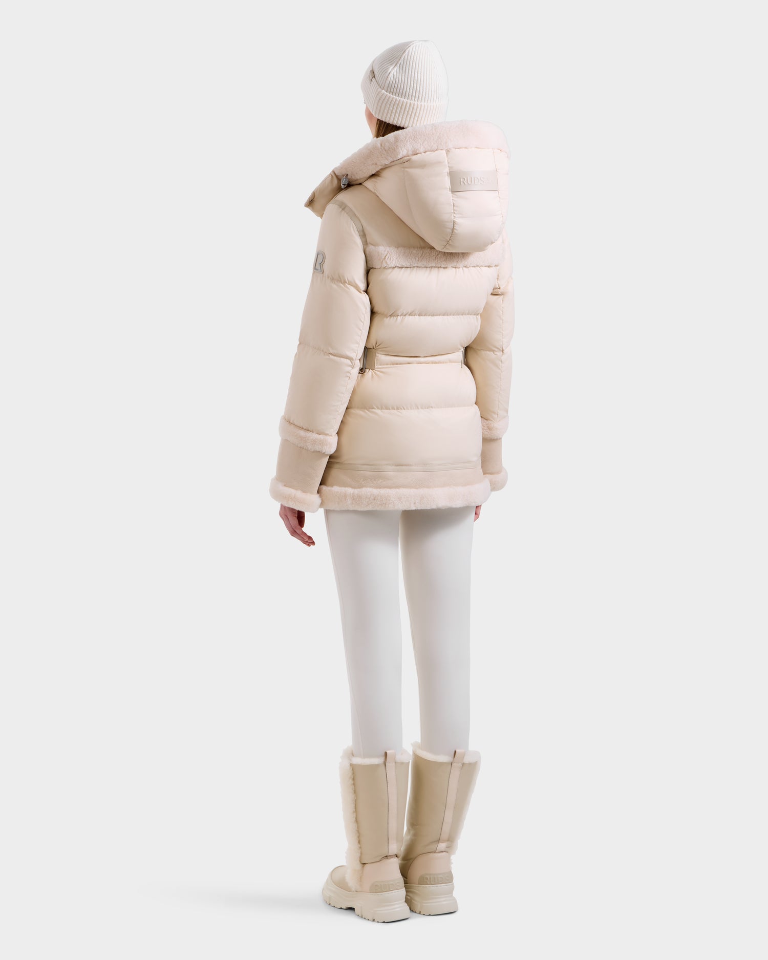 A person in the Rudsak TESSA - 8125615 Vanilla women’s down puffer jacket with a hood, matching leggings, winter boots, and a knit beanie stands facing away against a plain white background.