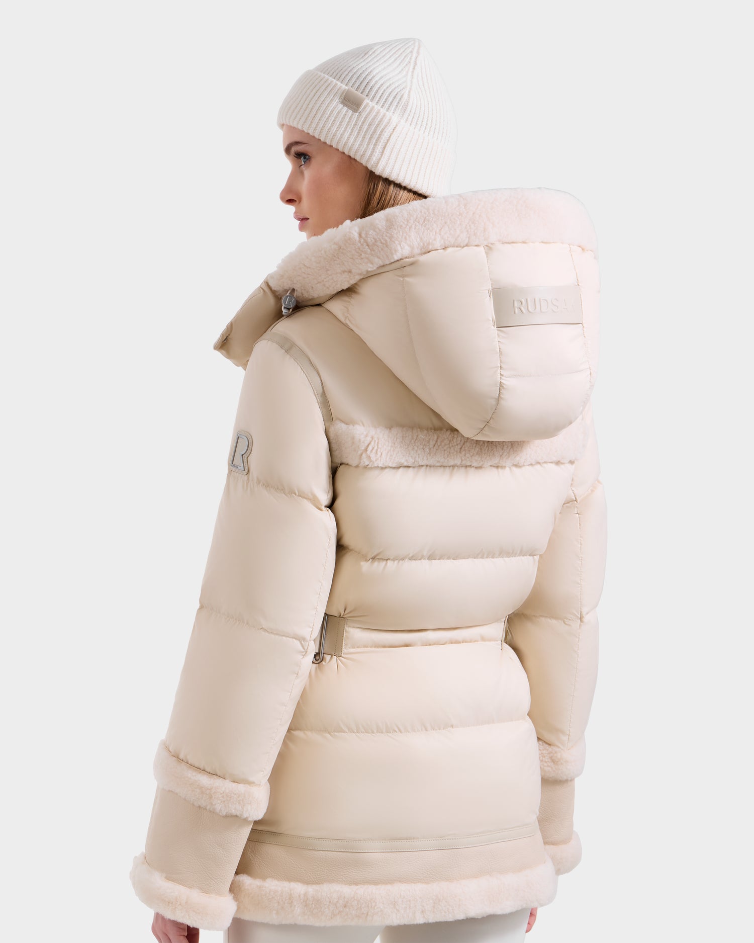 A woman wearing the Rudsak TESSA - 8125615 Vanilla down puffer jacket with a shearling trim hood and a matching knit beanie stands looking left against a plain white background.