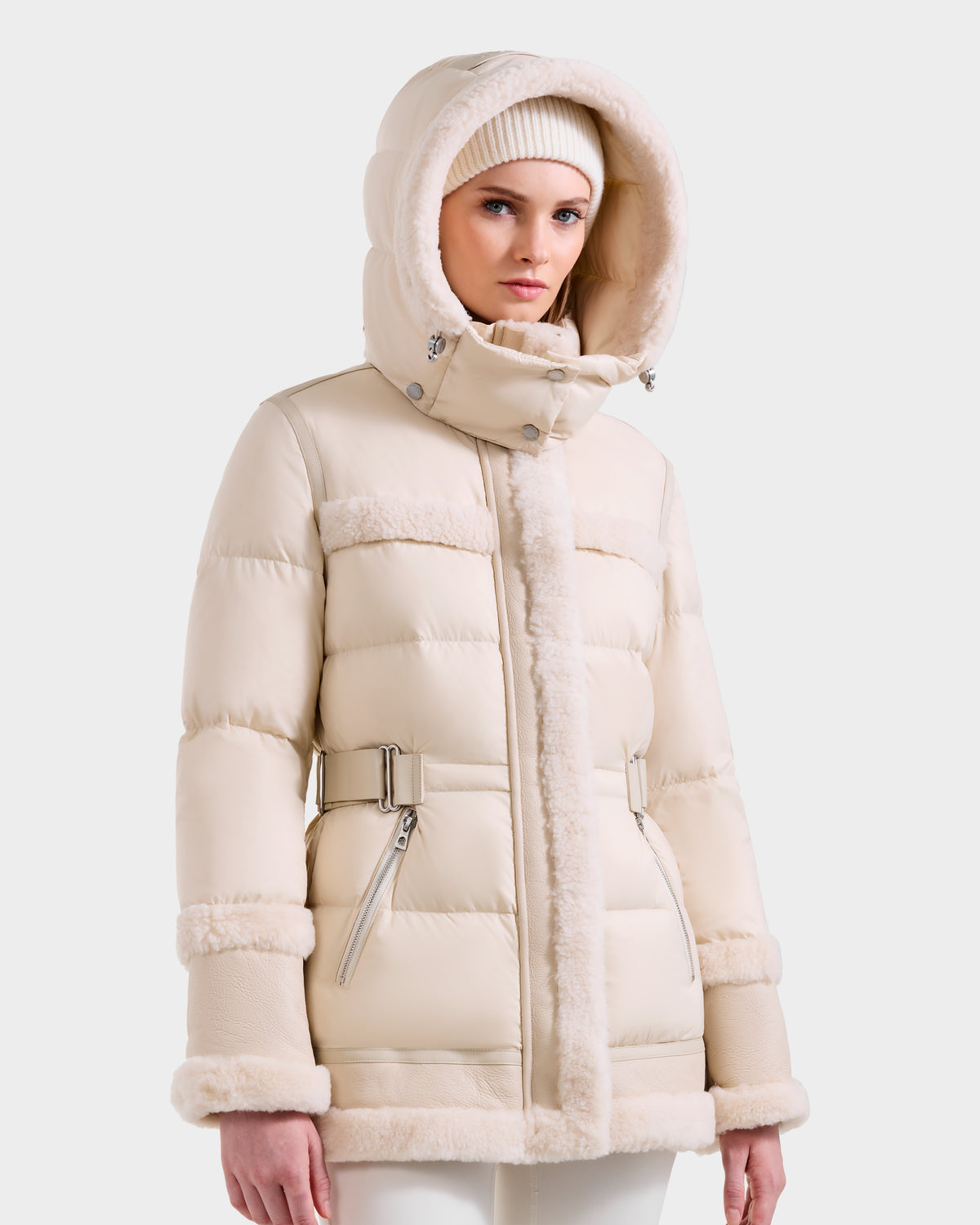 A woman in the Rudsak TESSA - 8125615 Vanilla down puffer jacket with shearling trim, belted sides, zippered pockets, and hood stands against a plain background, styled with a matching knit beanie.