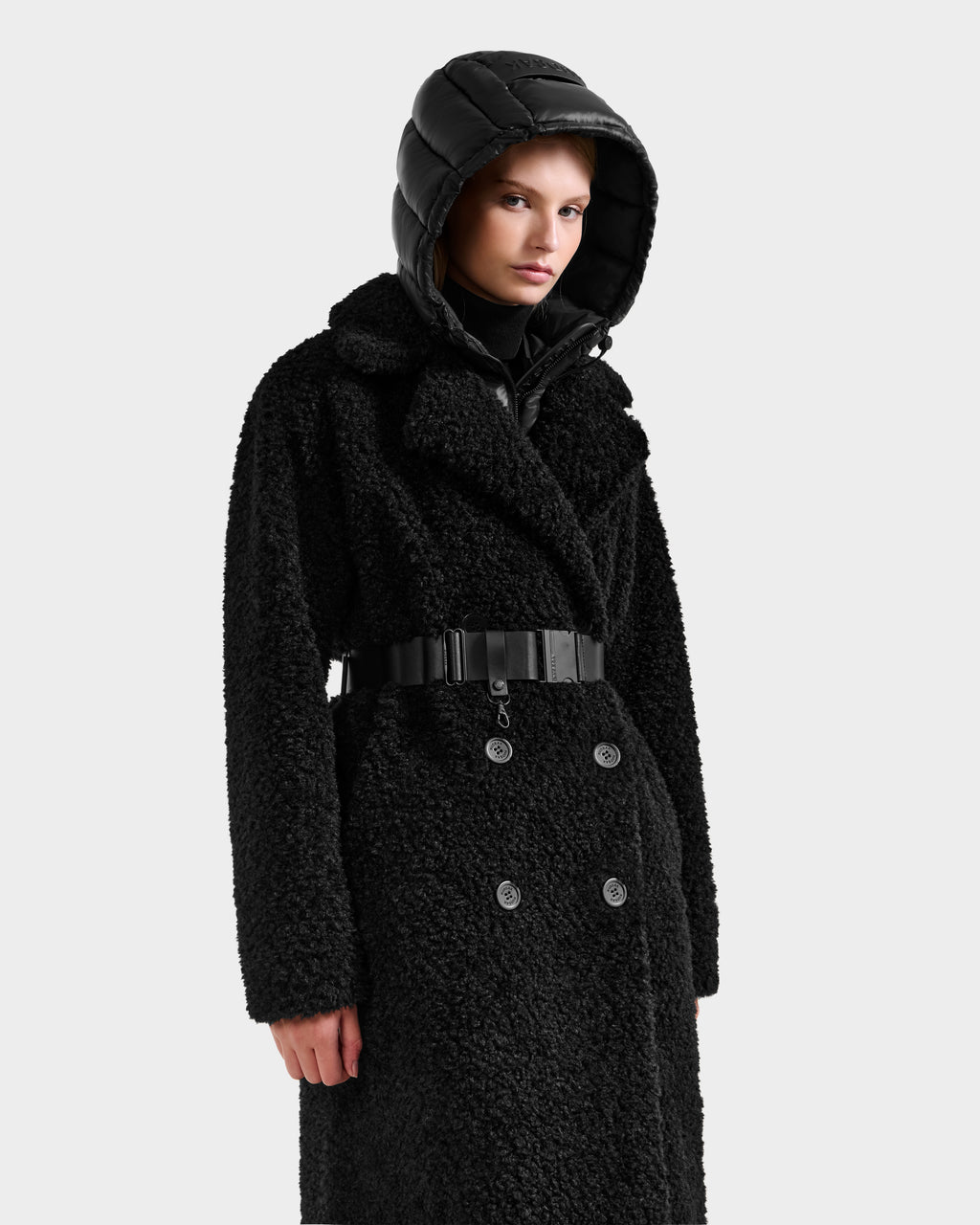A person wearing the Rudsak TINA - 8125968 Black faux fur teddy trench coat with a wide belt and shiny removable hooded bib, standing against a plain light background.