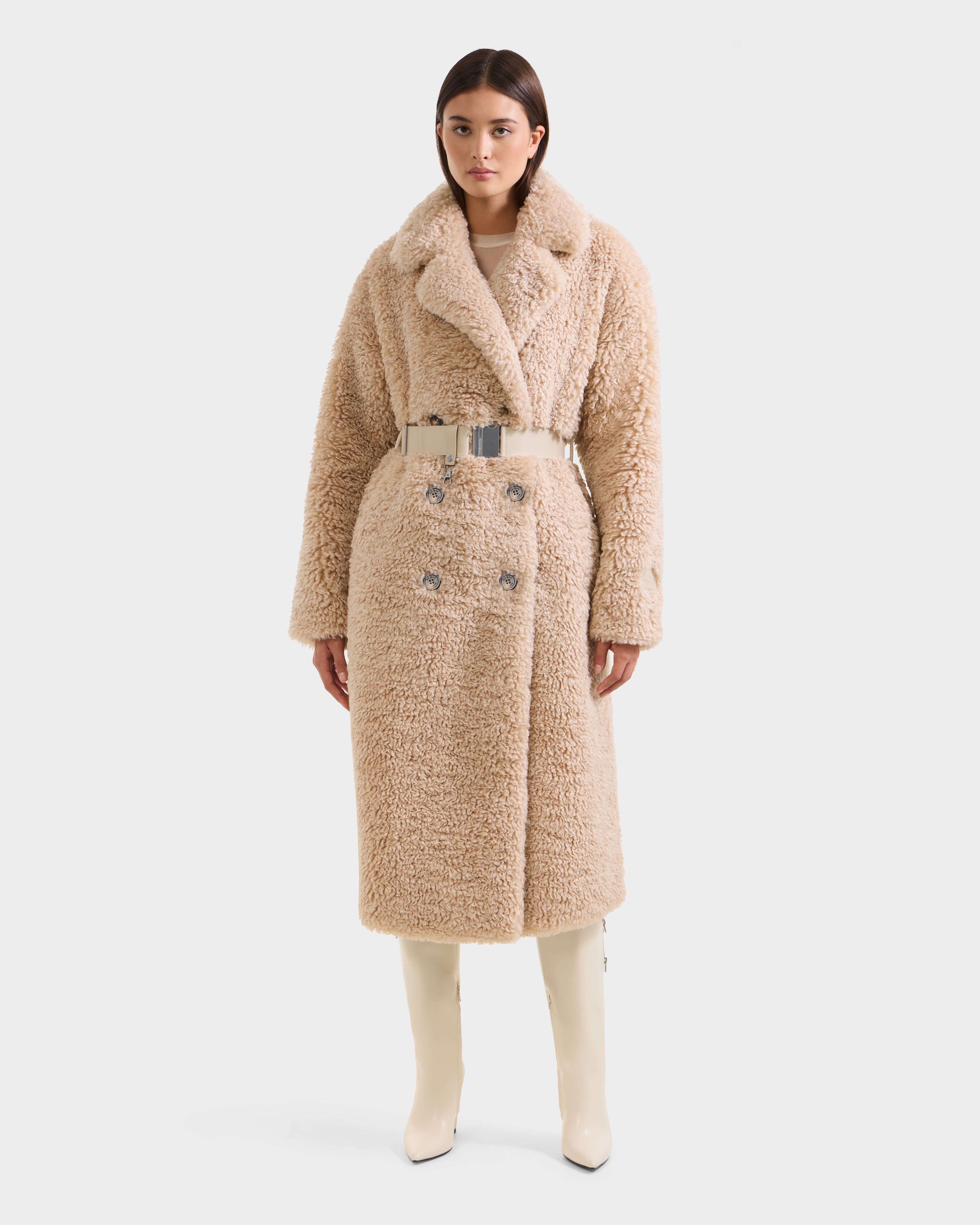 A woman models the Rudsak TINA - 8125968 Vanilla faux fur trench coat with a wide collar, decorative buttons, and metallic belt, paired with knee-high off-white boots against a plain white background.