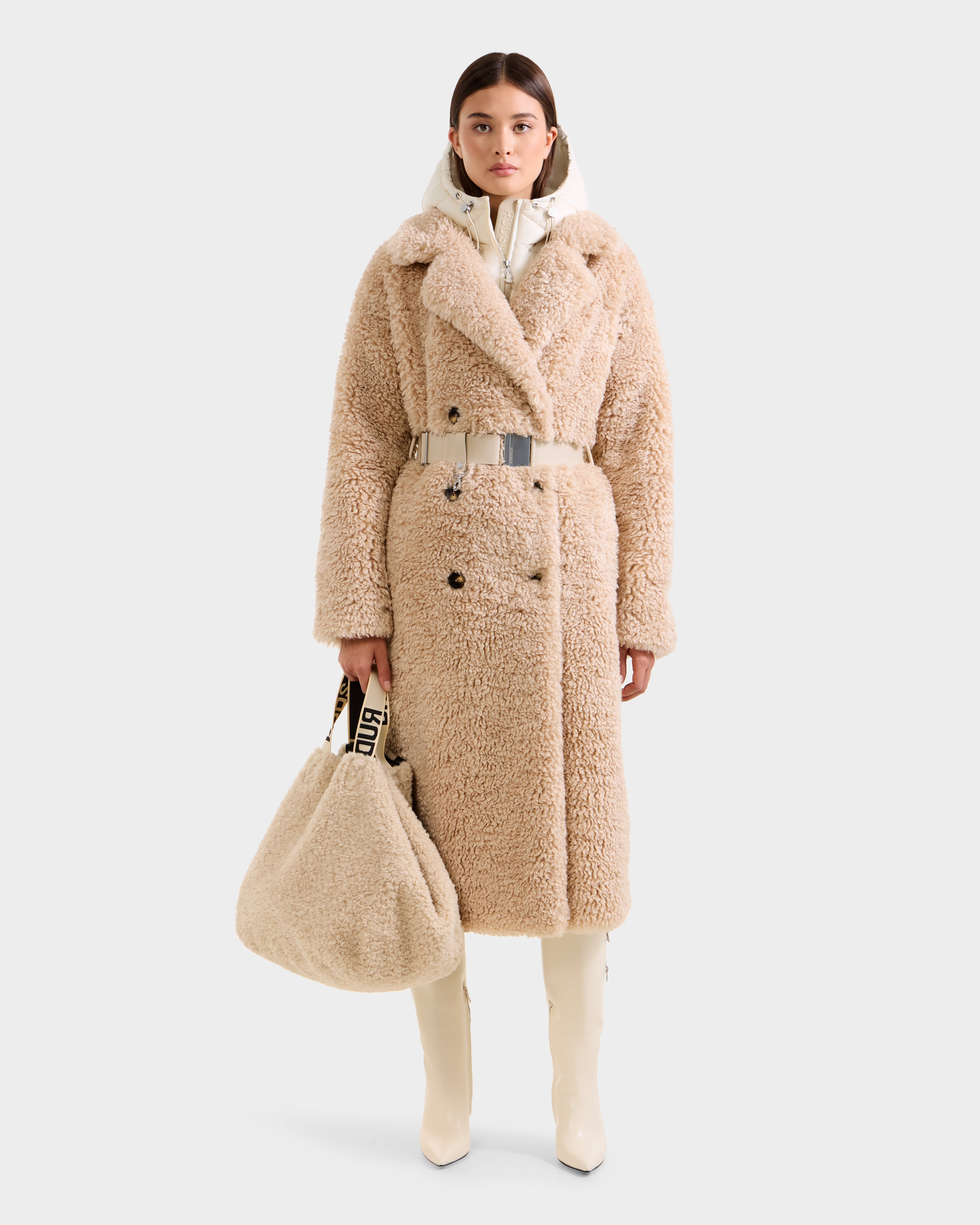 A woman faces forward in the Rudsak TINA - 8125968 Vanilla, a long beige faux fur trench coat with a belt, paired with cream high boots and a large matching faux fur bag with black-and-white strap, against a plain light background.