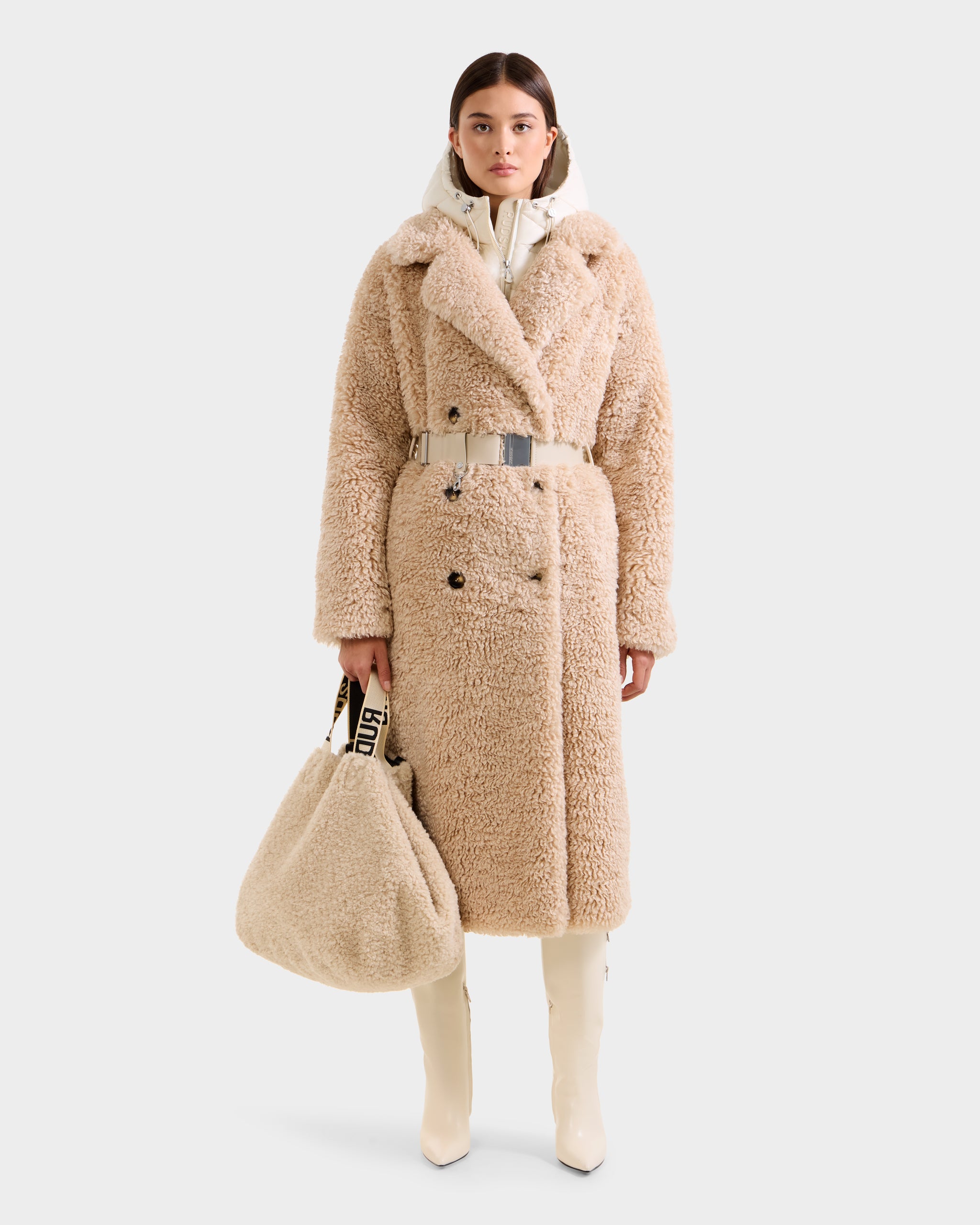 A woman faces forward in the Rudsak TINA - 8125968 Vanilla, a long beige faux fur trench coat with a belt, paired with cream high boots and a large matching faux fur bag with black-and-white strap, against a plain light background.