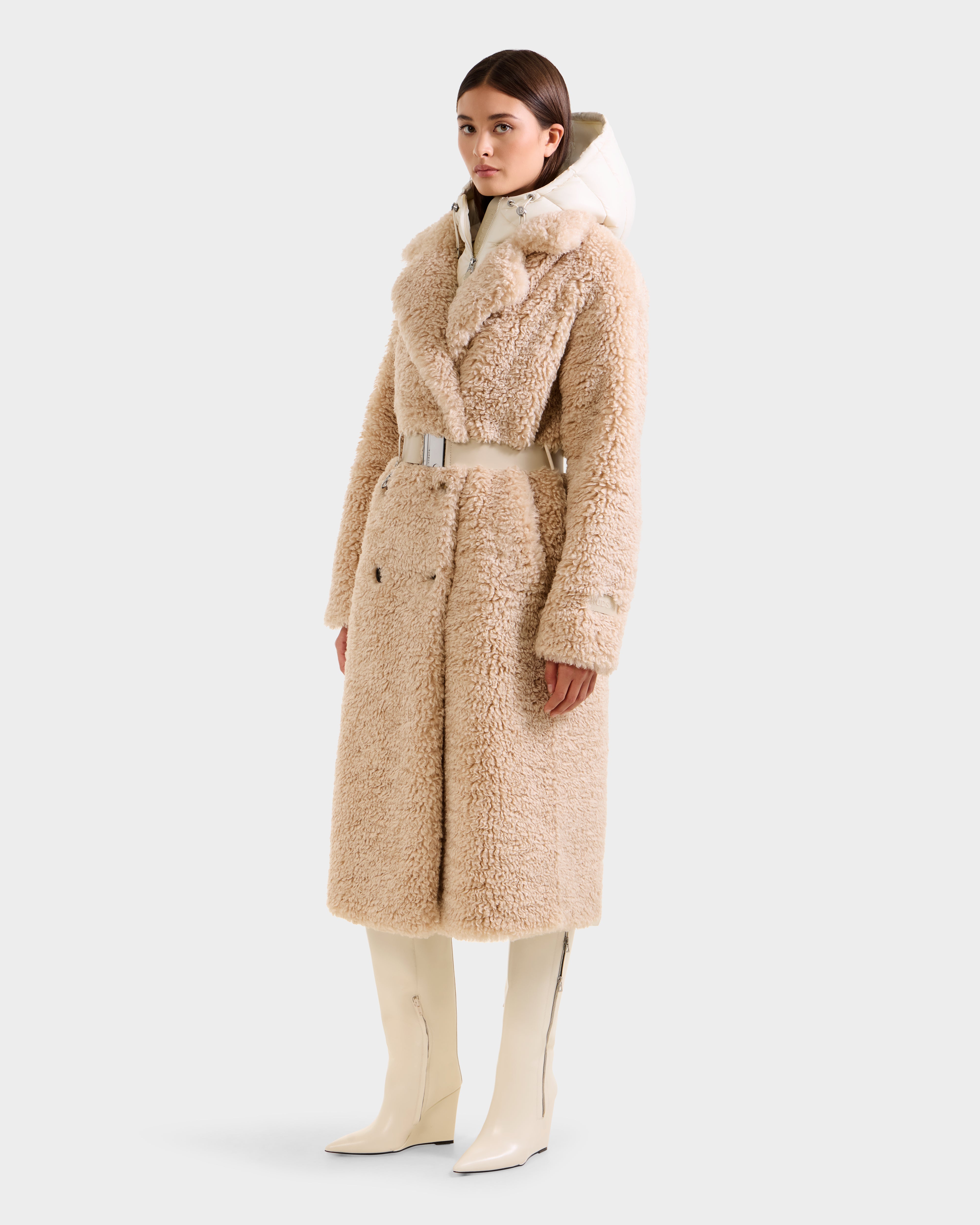 A woman wears the Rudsak TINA - 8125968 Vanilla faux fur trench coat with a wide belt, paired with a white turtleneck and knee-high white boots, standing against a plain light background.