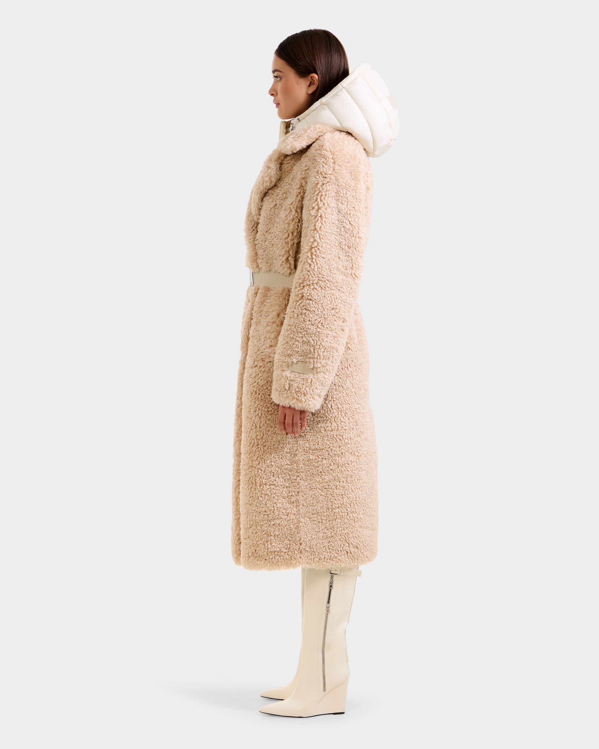 A woman in profile wears the Rudsak TINA - 8125968 Vanilla faux fur trench coat with a belted waist and removable hooded bib, paired with knee-high white boots, posed against a plain light background.