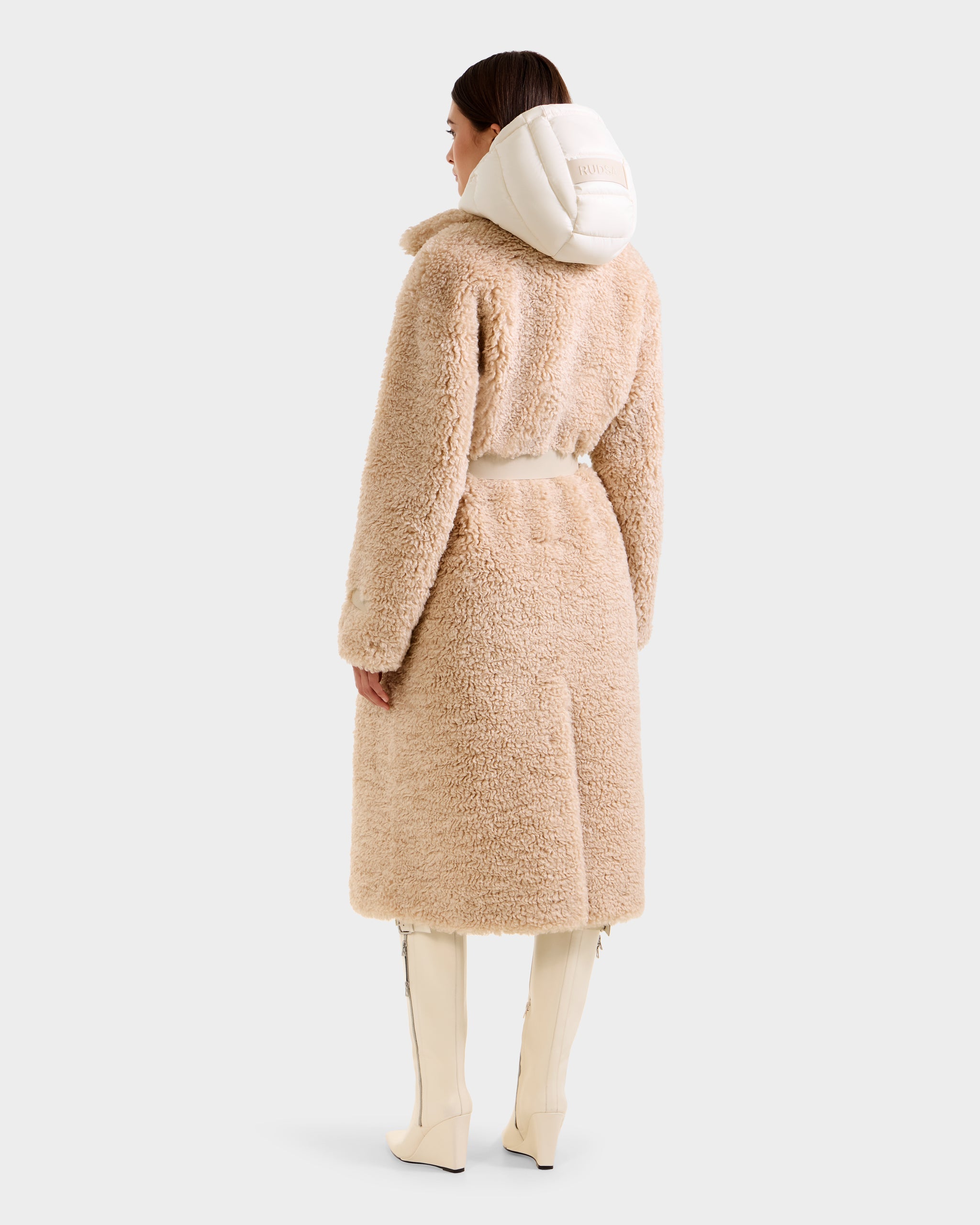 A woman faces away, wearing the Rudsak TINA - 8125968 Vanilla beige faux fur trench coat with a removable hooded bib, white knee-high boots, and her dark hair pulled back against a plain light background.