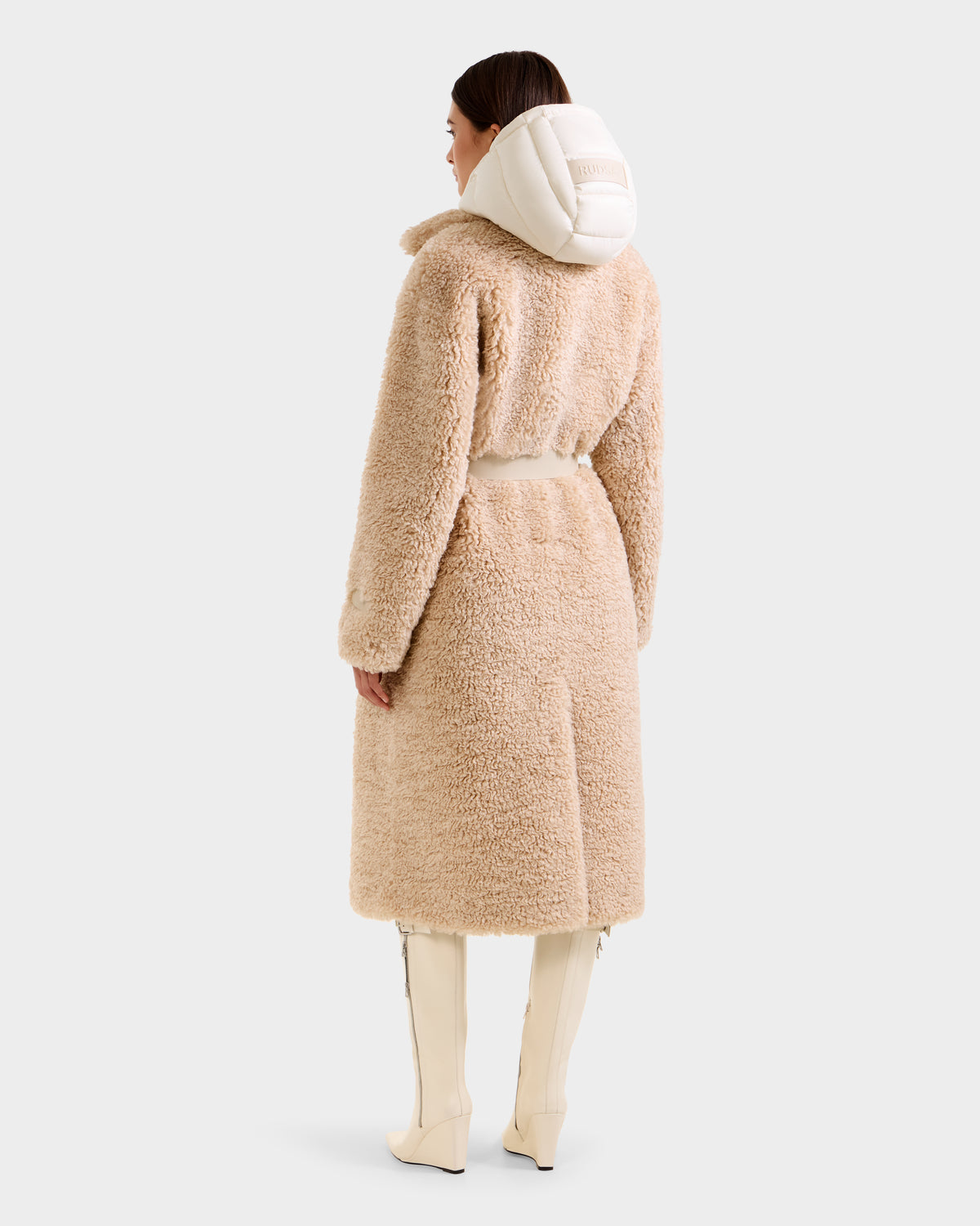 A woman faces away, wearing the Rudsak TINA - 8125968 Vanilla beige faux fur trench coat with a removable hooded bib, white knee-high boots, and her dark hair pulled back against a plain light background.