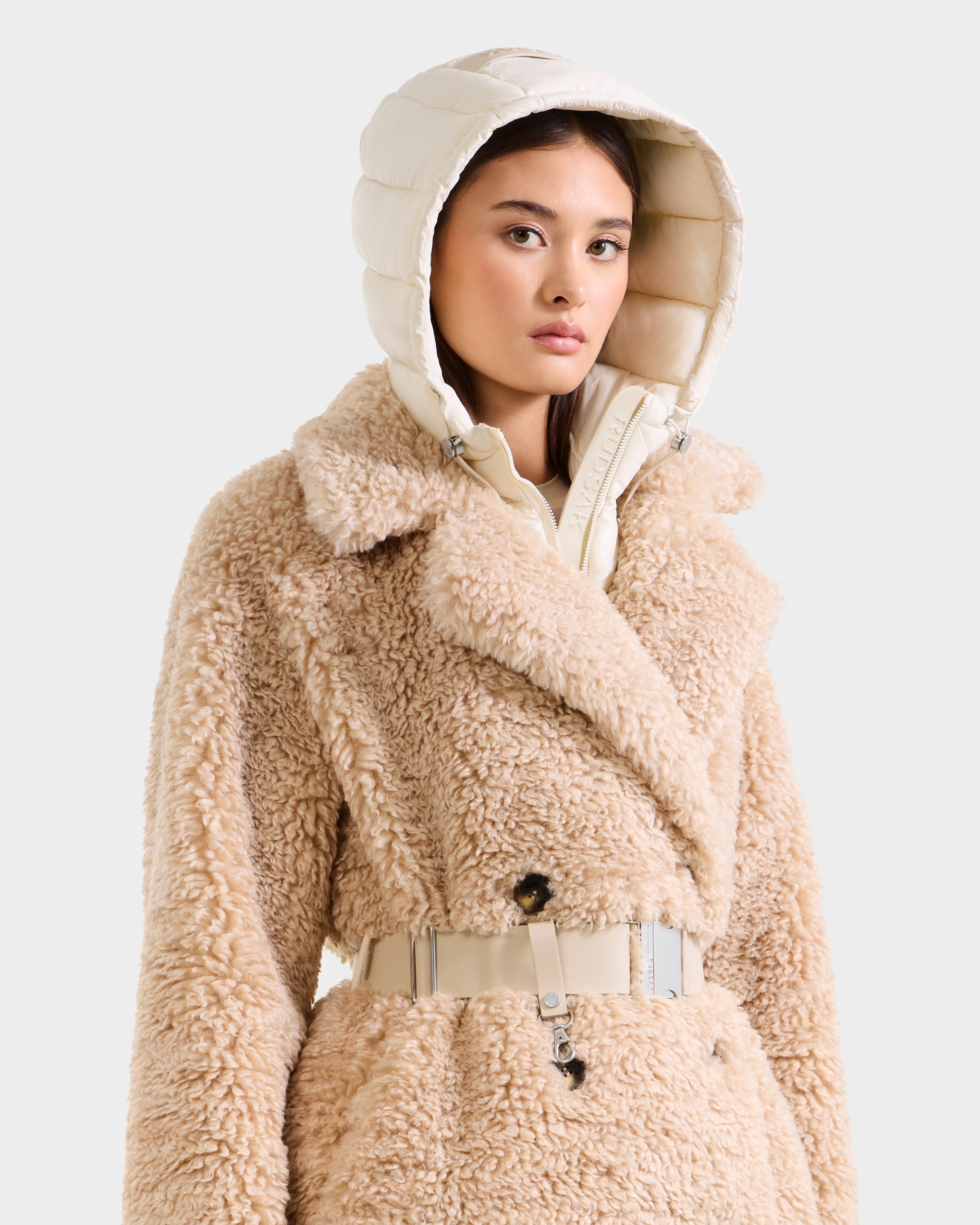 A woman wears the Rudsak TINA - 8125968 Vanilla trench coat, featuring beige faux fur, a wide collar, and a light cream removable hooded bib, standing against a plain light background.