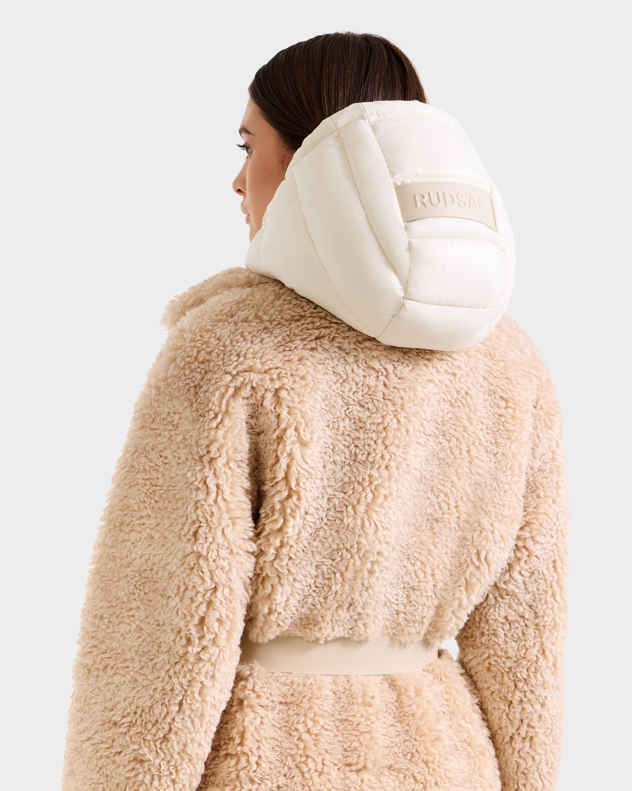 A person is shown from the back wearing Rudsak's TINA - 8125968 Vanilla, a beige teddy trench coat with a cream padded removable hooded bib featuring a RUDSAK patch, against a plain background.