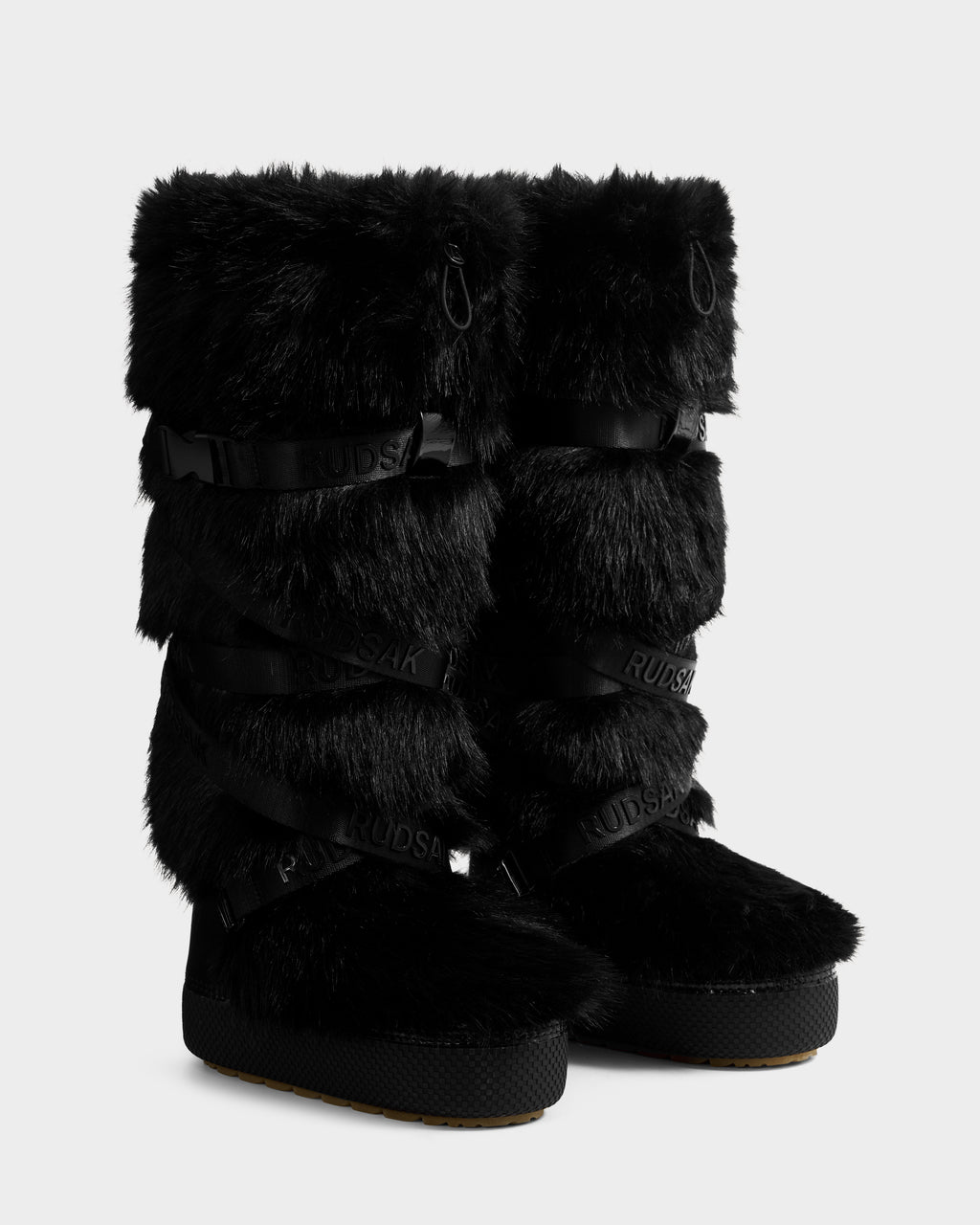 The Rudsak TORIN - 8225029 Black women's faux fur moon boots feature thick rubber soles and crisscrossing black straps with the Rudsak logo, making them perfect for winter style, shown on a plain white background.