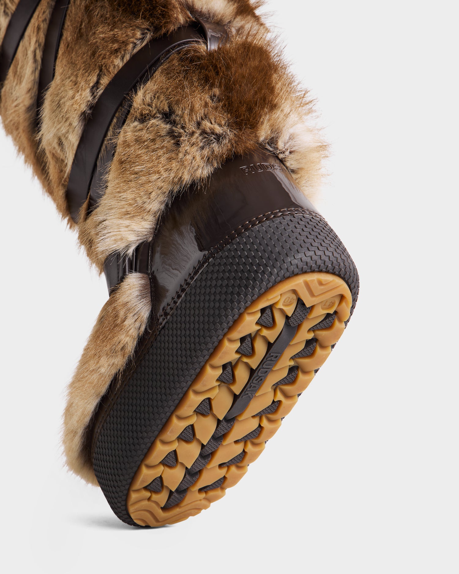 A close-up of Rudsak's TORIN - 8225029 Mix Mocha women’s moon boot highlights its faux fur, textured rubber sole with detailed tread, and glossy dark brown accents for a chic winter look, showcasing the bottom and side materials.