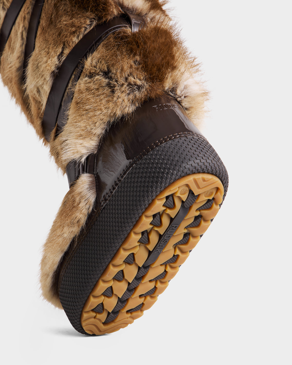 A close-up of Rudsak's TORIN - 8225029 Mix Mocha women’s moon boot highlights its faux fur, textured rubber sole with detailed tread, and glossy dark brown accents for a chic winter look, showcasing the bottom and side materials.