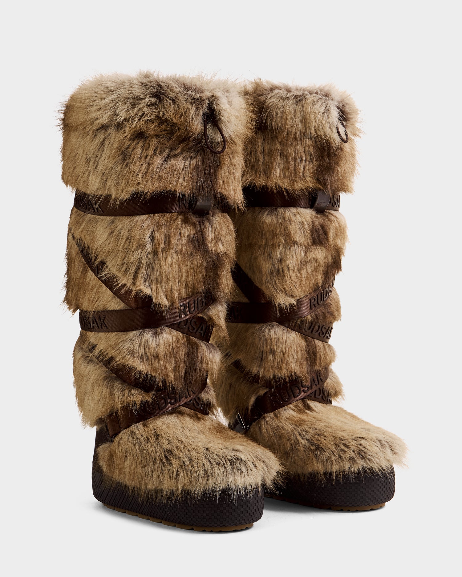 A pair of tall women’s Rudsak TORIN - 8225029 Mix Mocha faux fur moon boots in brown, with black soles and dark crisscrossed straps. These winter boots have a bulky, layered look and textured faux fur covering most surfaces.