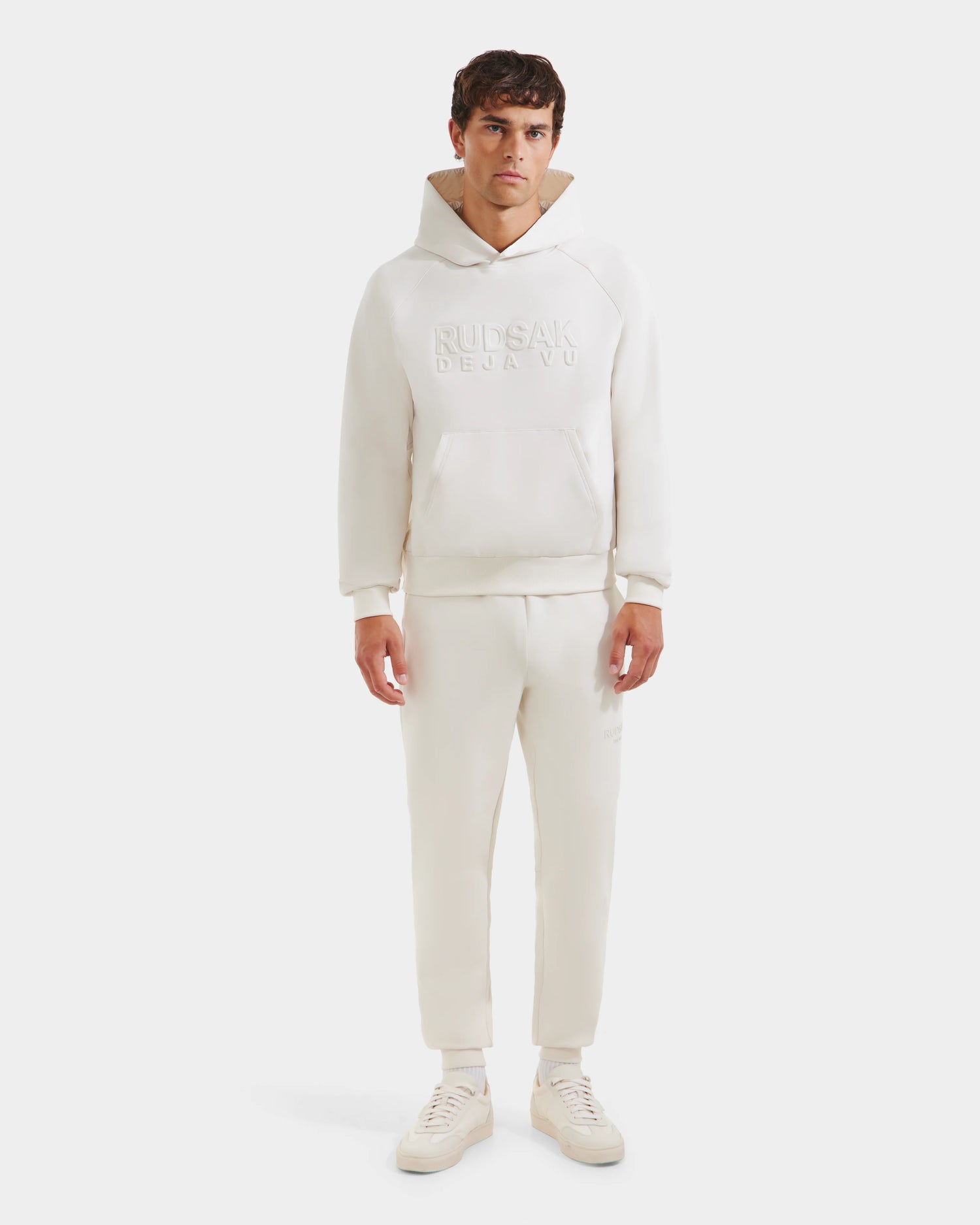 A man stands against a white background wearing the Rudsak TREVIS - 6525599 Vanilla mens cotton-blend hoodie and jogger set with RUDSAK DEJA VUE embossed on the hoodie, paired with off-white sneakers.