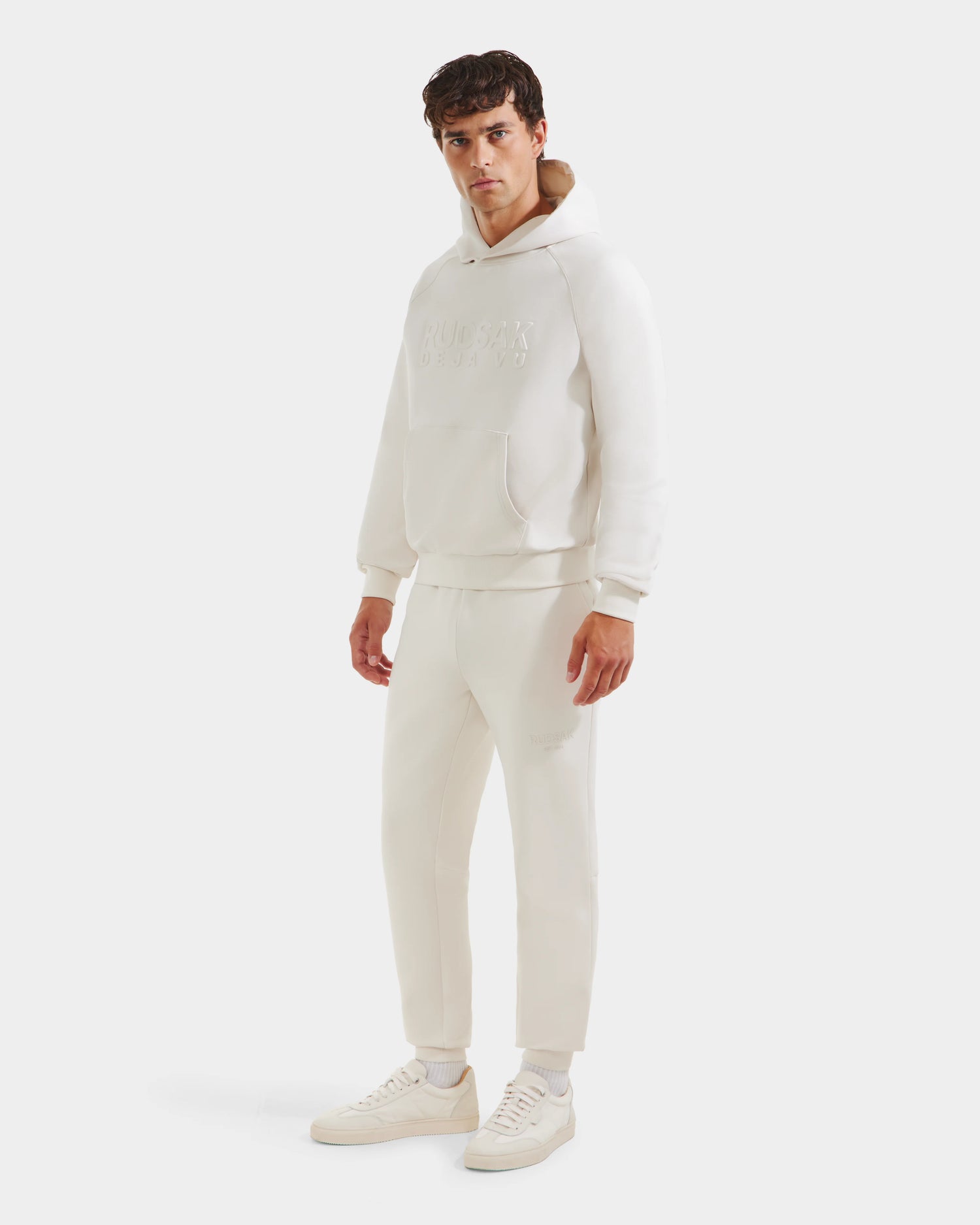 A man faces forward in a Rudsak TREVIS - 6525599 Vanilla men’s cotton-blend hoodie with front pocket and embossed text, matching joggers, and white sneakers. The background is plain white.