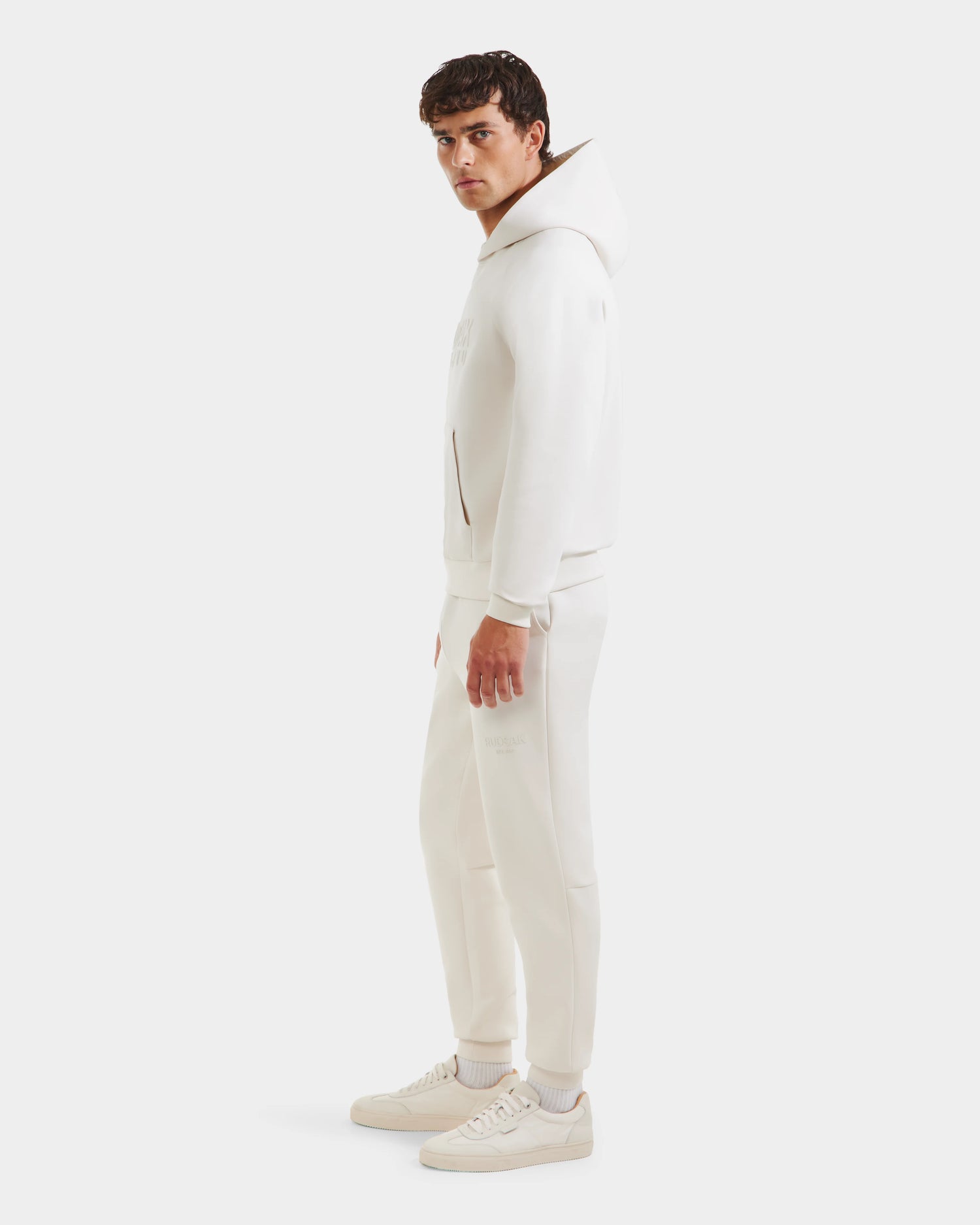 A man stands sideways in Rudsaks TREVIS - 6525599 Vanilla cotton-blend hoodie and joggers, socks, and sneakers against a white background, looking at the camera with a neutral expression that reflects Rudsaks clean style.