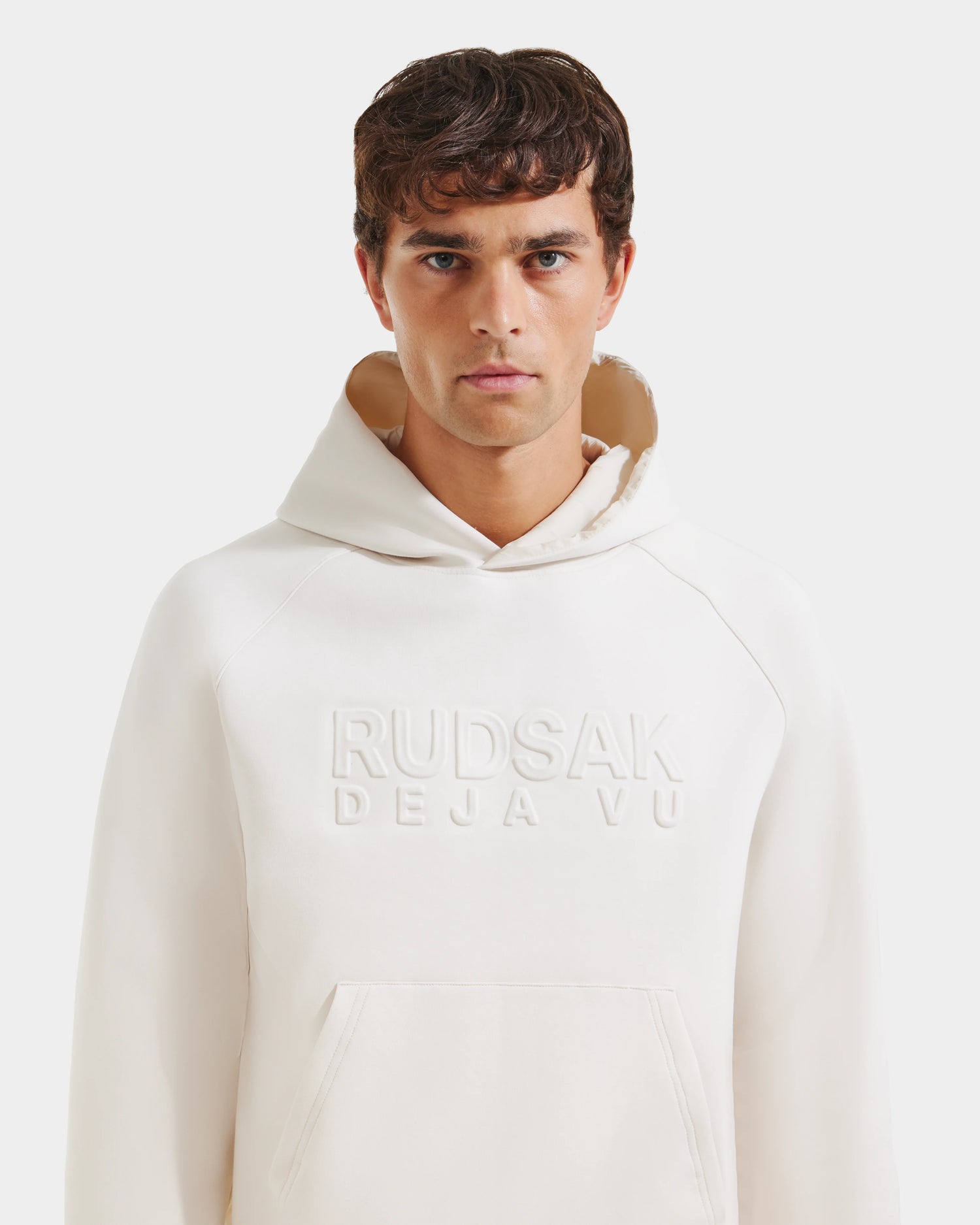 A man with short brown hair wears the Rudsak TREVIS - 6525599 Vanilla men’s cotton-blend hoodie with RUDSAK DEJA VUE embossed on the front, standing against a plain white background and looking directly at the camera.