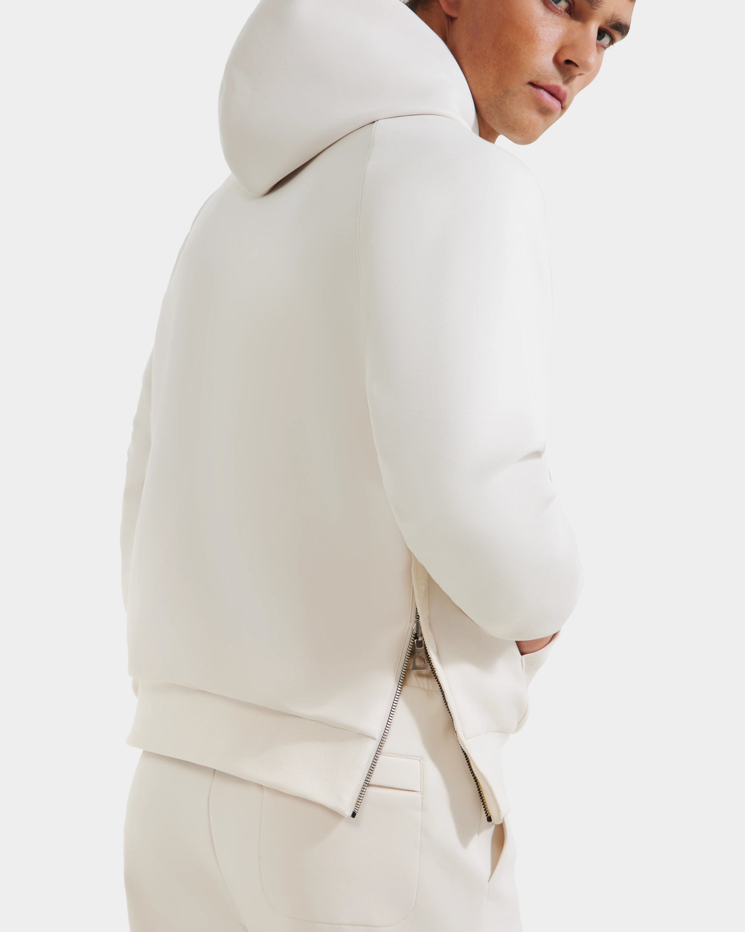 A person wearing the Rudsak TREVIS - 6525599 Vanilla hoodie and matching pants stands slightly turned, highlighting the hoodie’s sleek zippered detail against a plain white background.