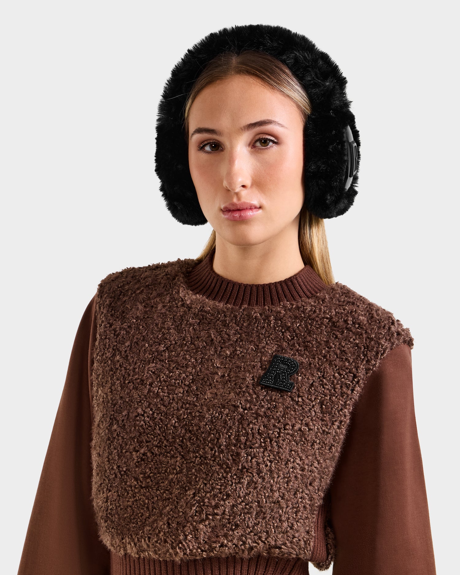 A woman with long blonde hair wears black Rudsak VALENCIA - 8425543 earmuffs and a textured brown sweater with a black R patch, standing against a plain light background.