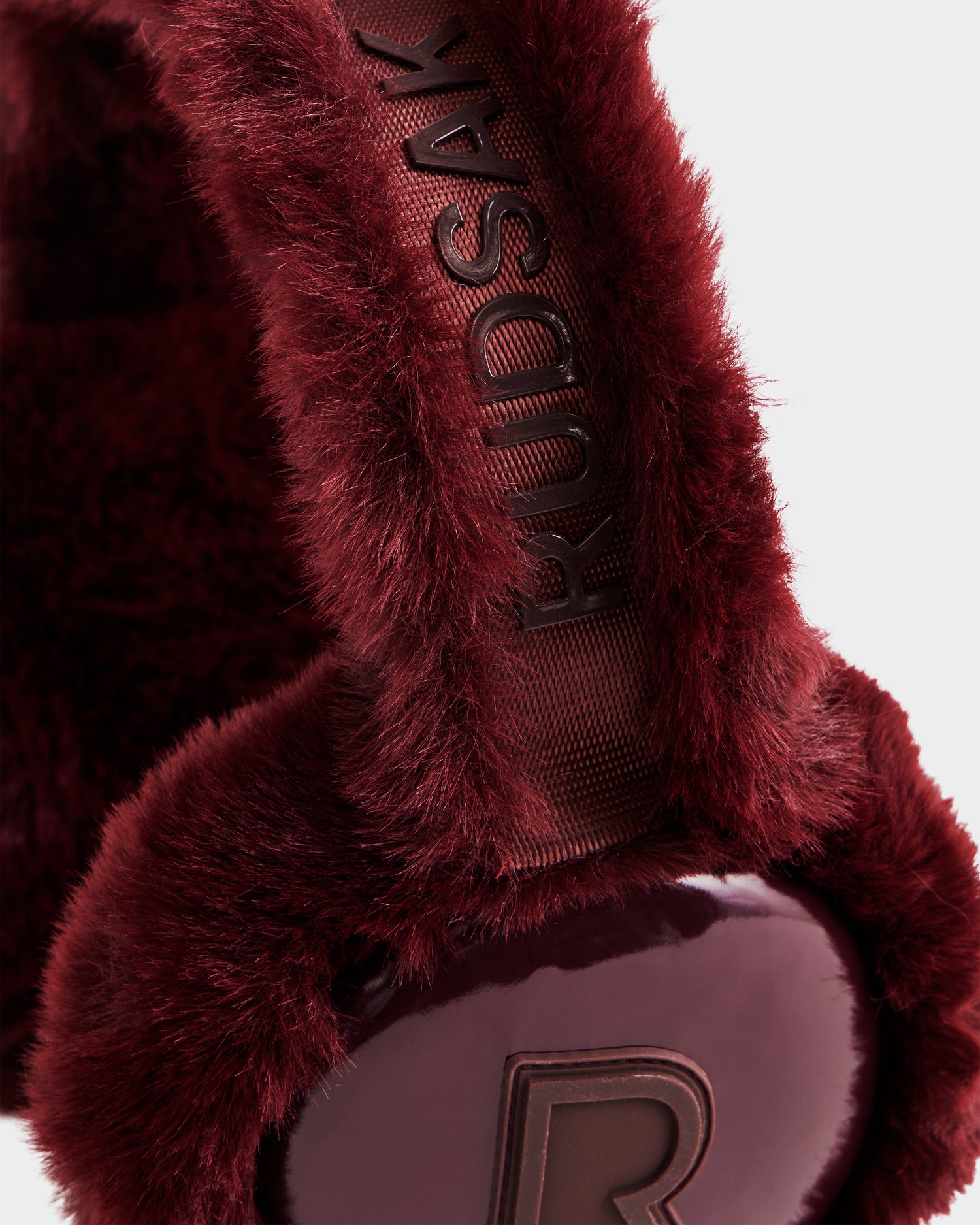 Close-up of Rudsak VALENCIA - 8425543 Bordeaux earmuffs, a chic winter accessory with burgundy faux fur, the brand name on the band, and a shiny R logo on the ear cover.