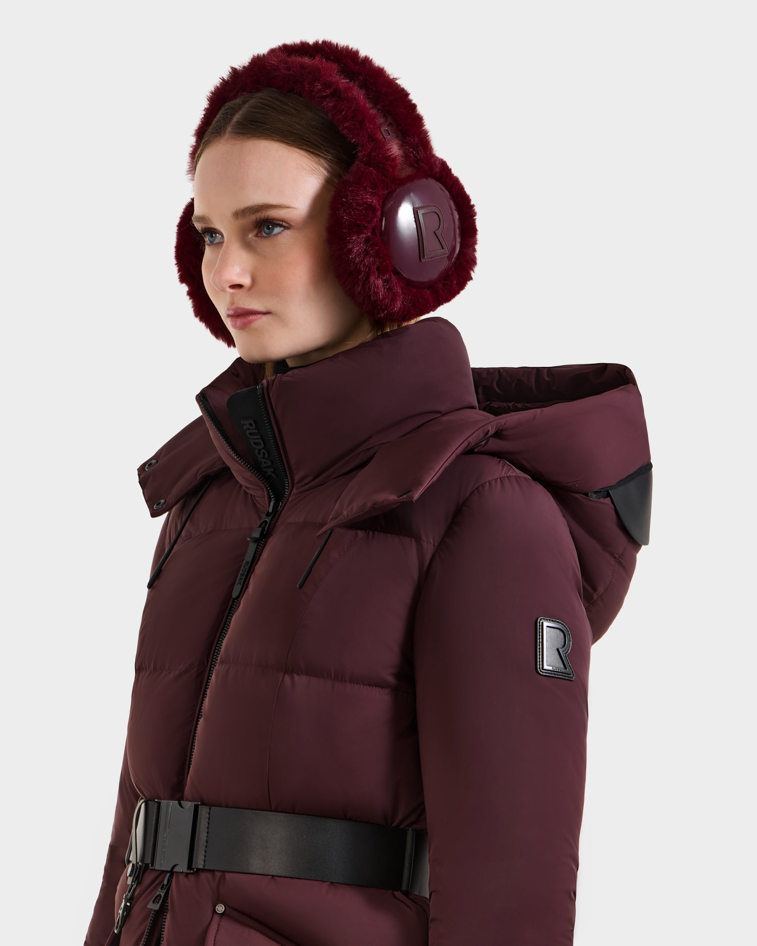 A woman wearing the Rudsak VALENCIA - 8425543 Bordeaux puffer jacket with a hood, black belt, and sleeve patch "R," styled with faux fur earmuffs, looks to the side—making this winter piece a standout accessory.